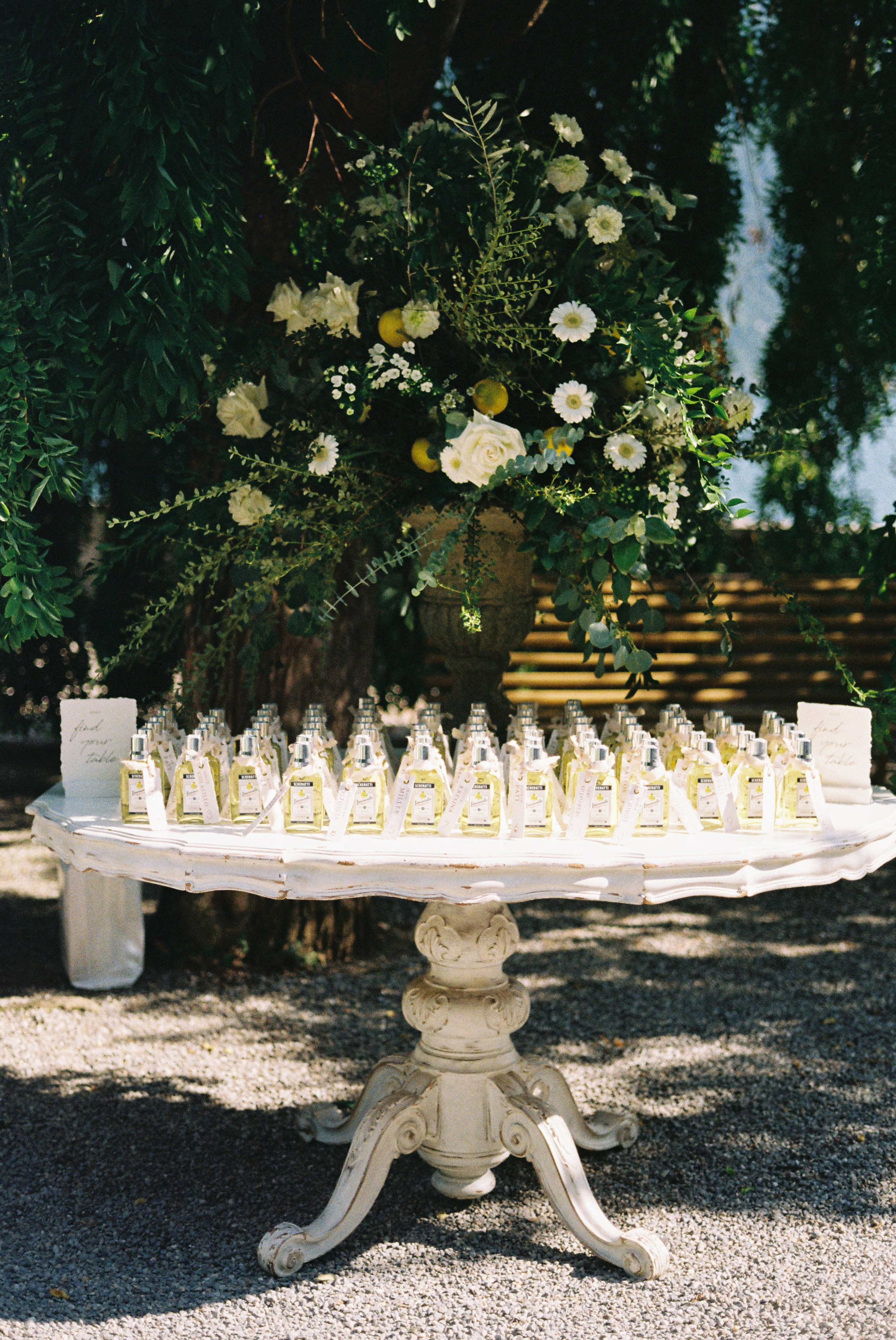 Decorative table with personalized wedding favors, set under a tree with a floral arrangement.