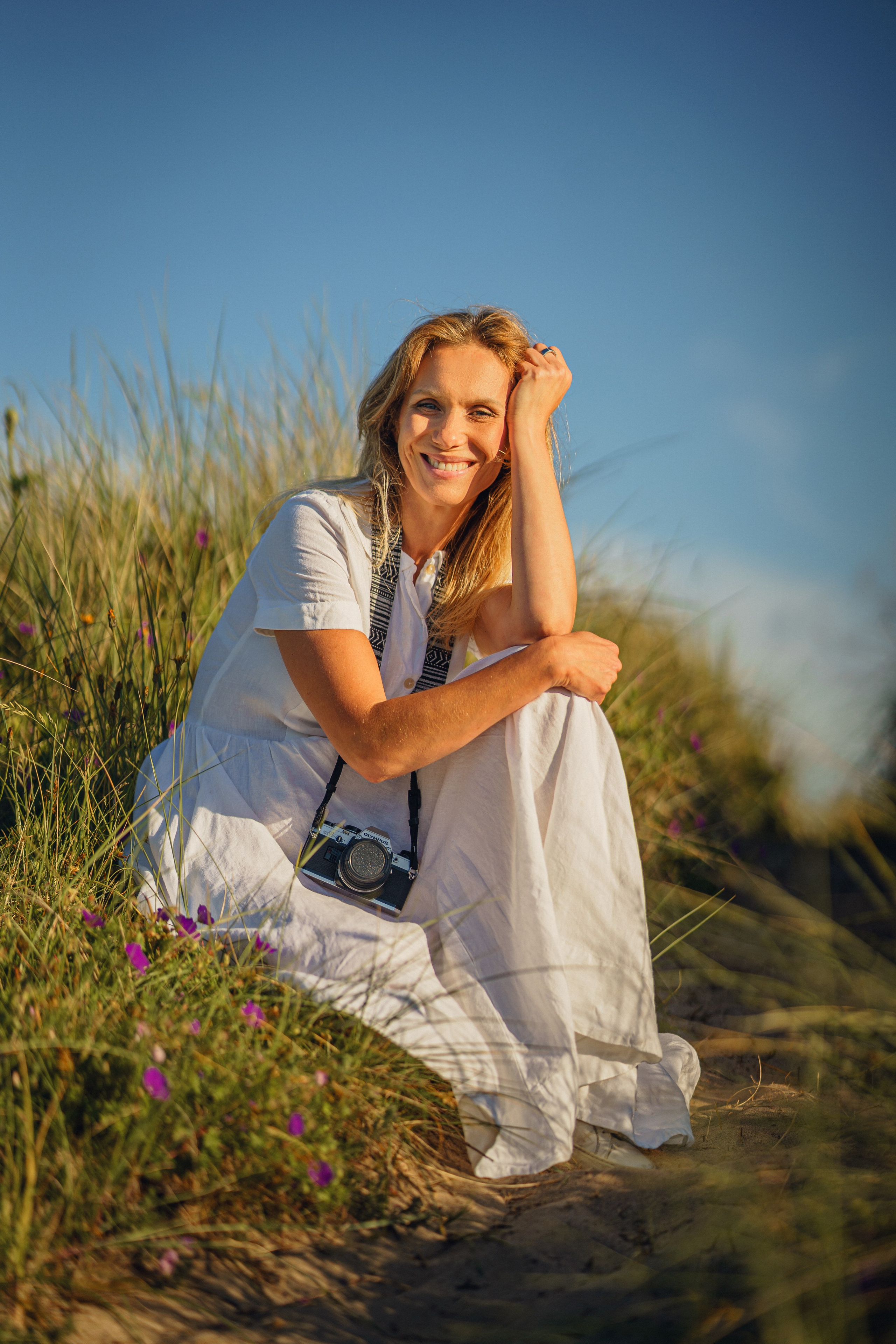 A beautiful, very happy woman enjoys the rays of the sunset while sitting in the grass by the seashore with a vintage camera.