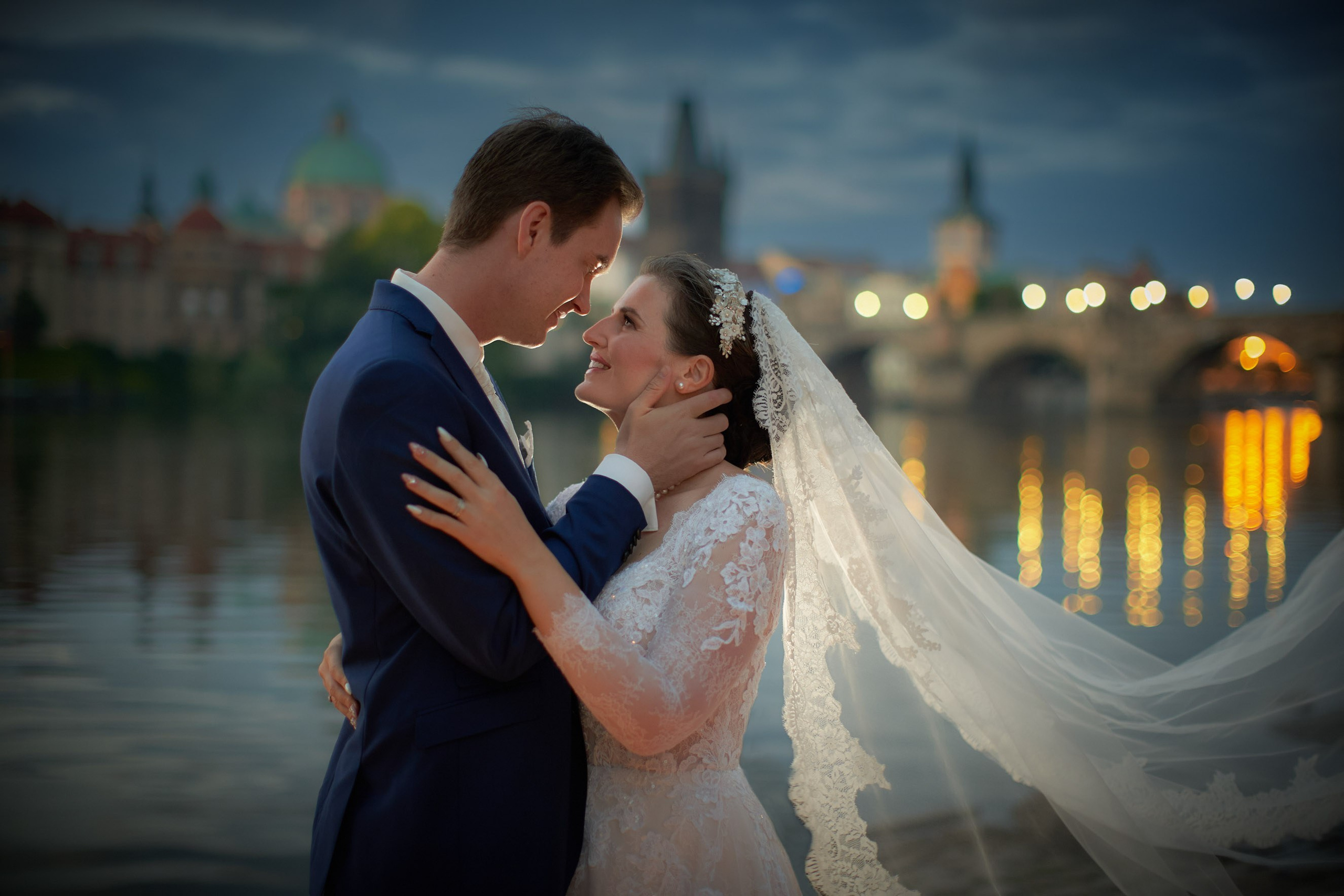 Groom tenderly touching bride's face near the Vltava River, with the illuminated Charles Bridge reflecting in the water.