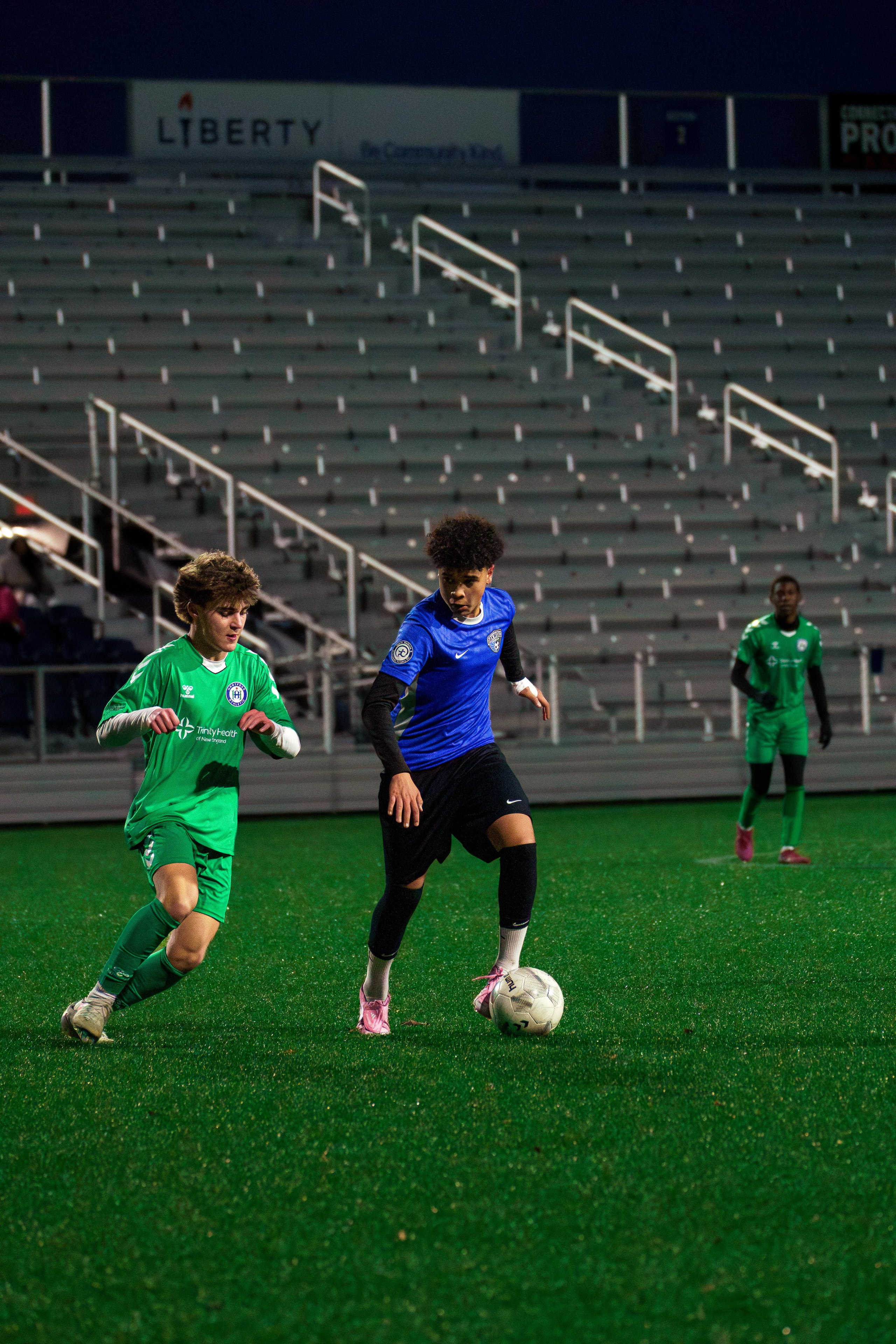 Soccer player dribbling forward under pressure as an opposing defender rushes in with arms raised to challenge for the ball.