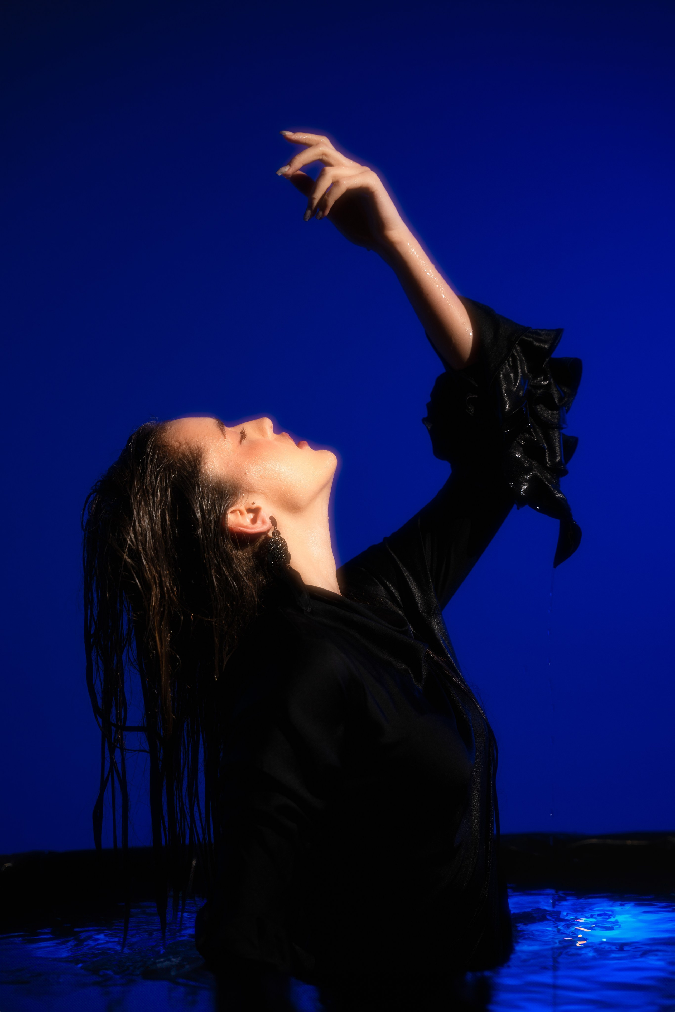 Editorial portrait of a woman reaching upward against a deep blue backdrop, photographed with dramatic color and a fashion-led composition.