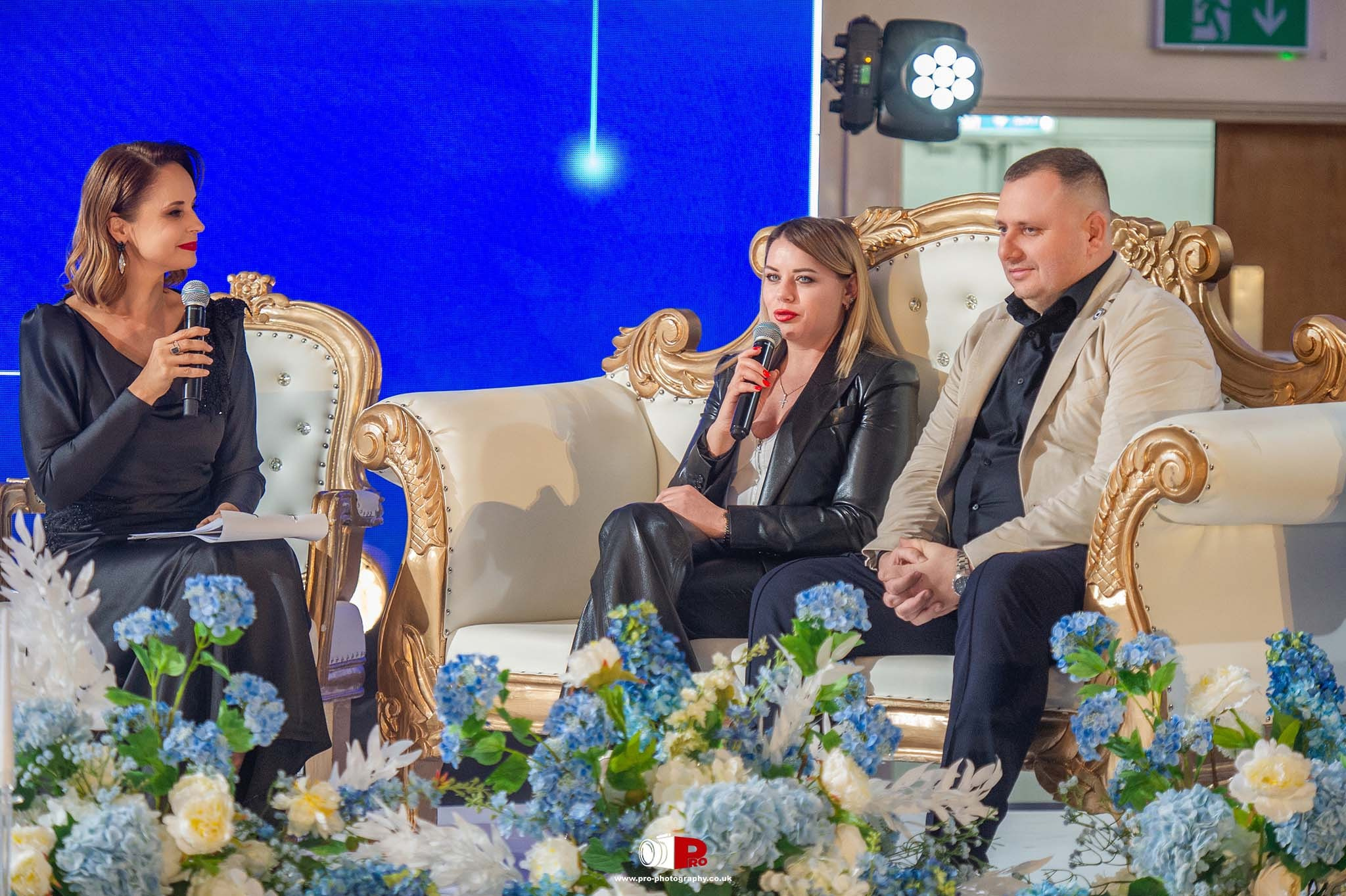 A panel discussion with a woman speaker holding a microphone was followed by two seated individuals on a stage adorned with decorative flowers.