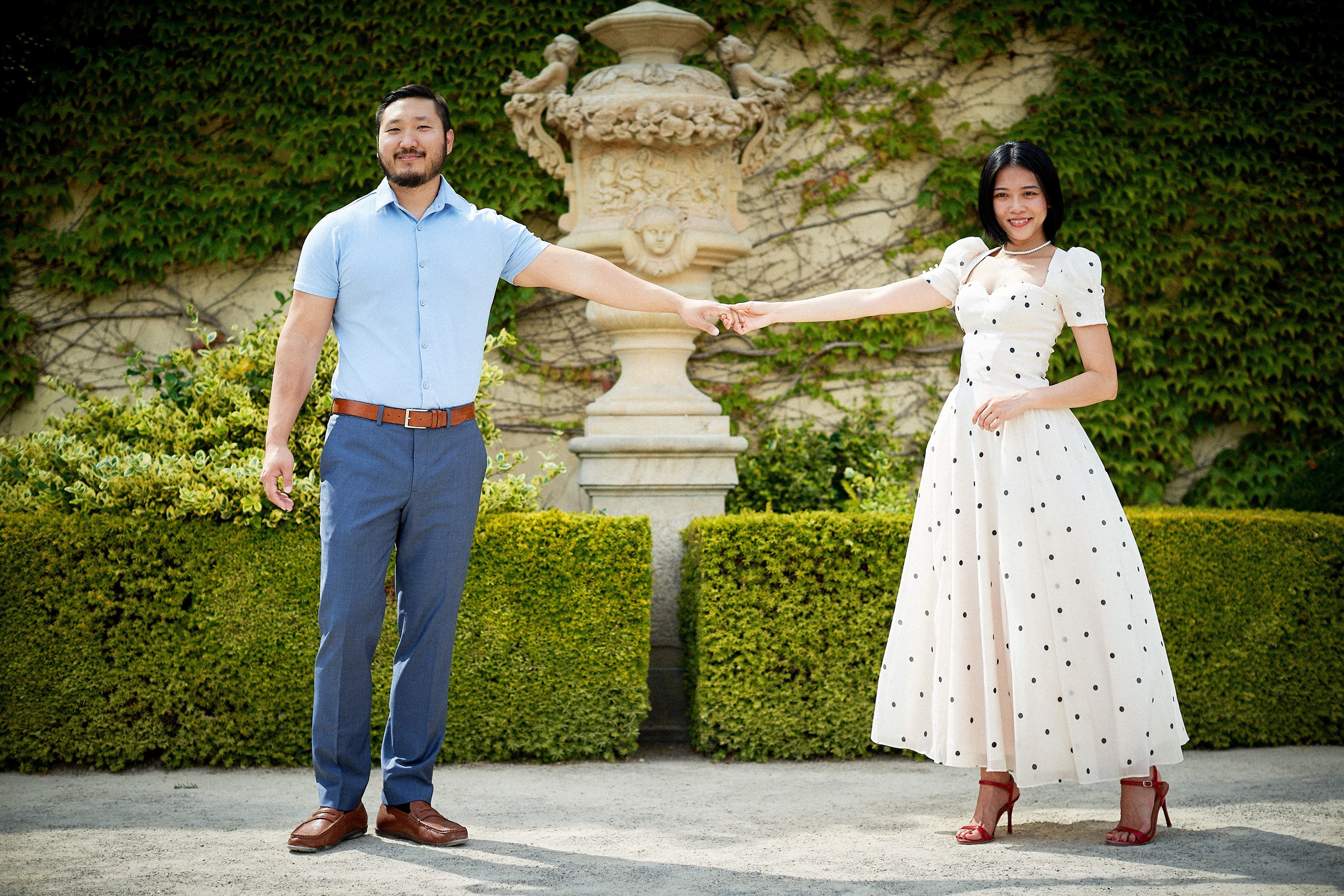 Engaged couple holding hands for an impromptu dance beside ivy-covered wall in Vrtba Garden.