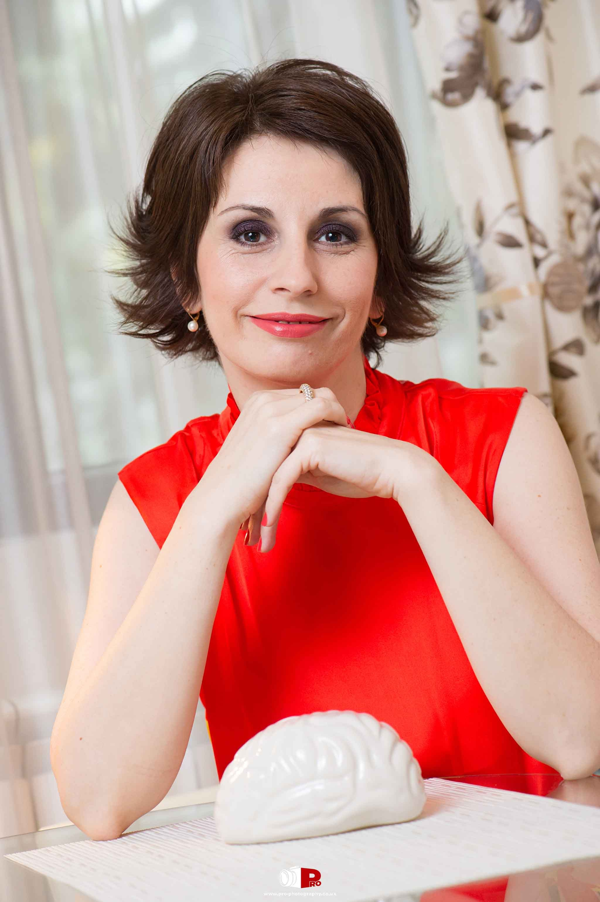 Professional portrait of a woman in a vibrant red dress, resting her hands on a table with a ceramic brain sculpture, smiling warmly.