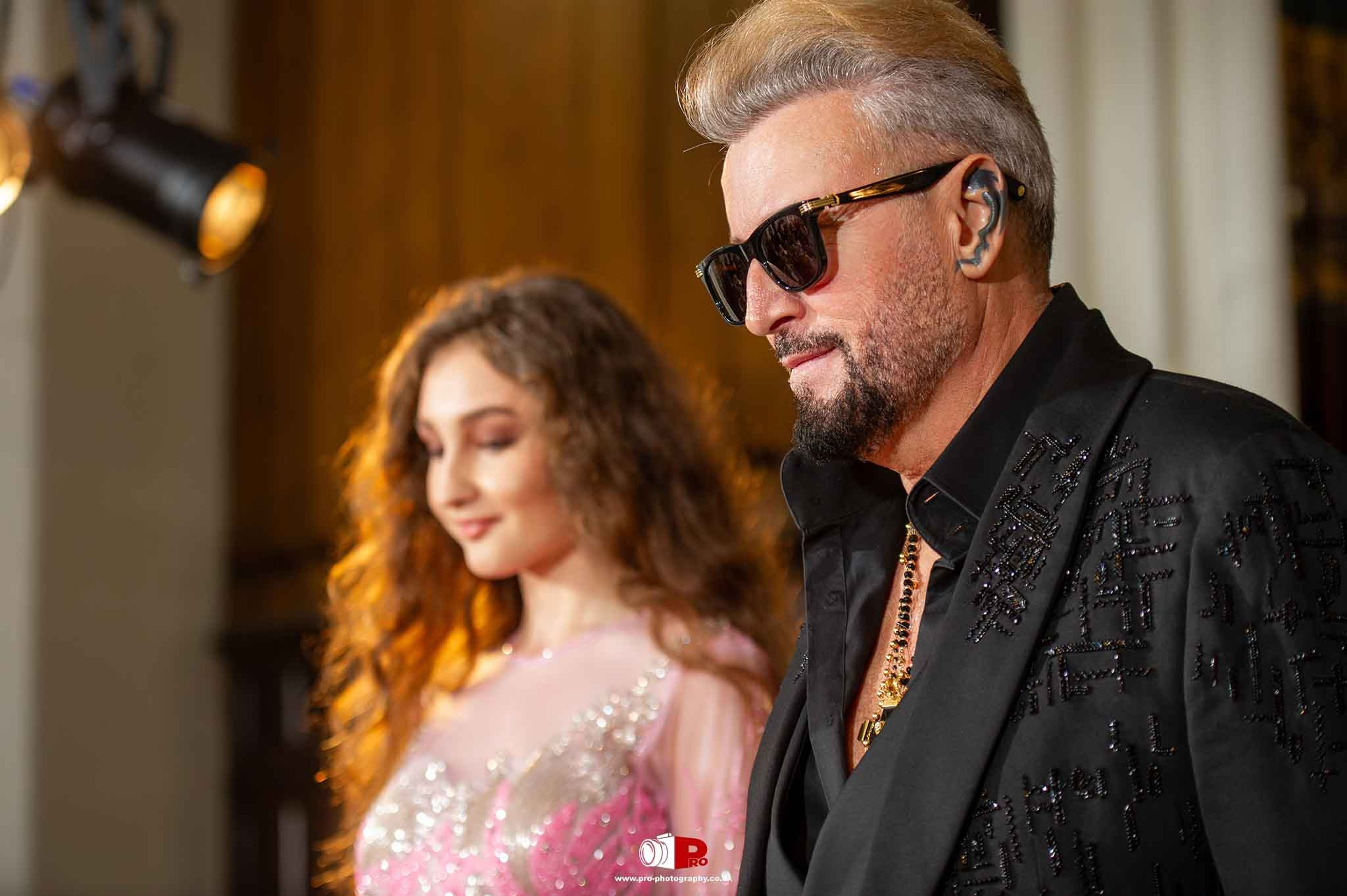 A renowned designer, Catalin Botezatu, in a black embroidered suit, walking down the runway with a model in a glittering pink dress.
