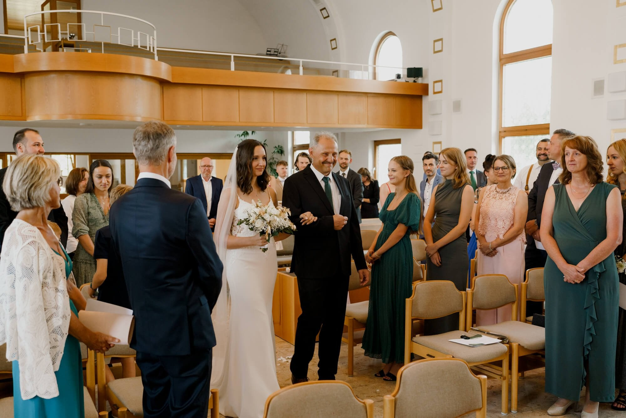 Bride walking down the aisle with her bouquet as guests stand and look on in a light-filled Stuttgart ceremony hall