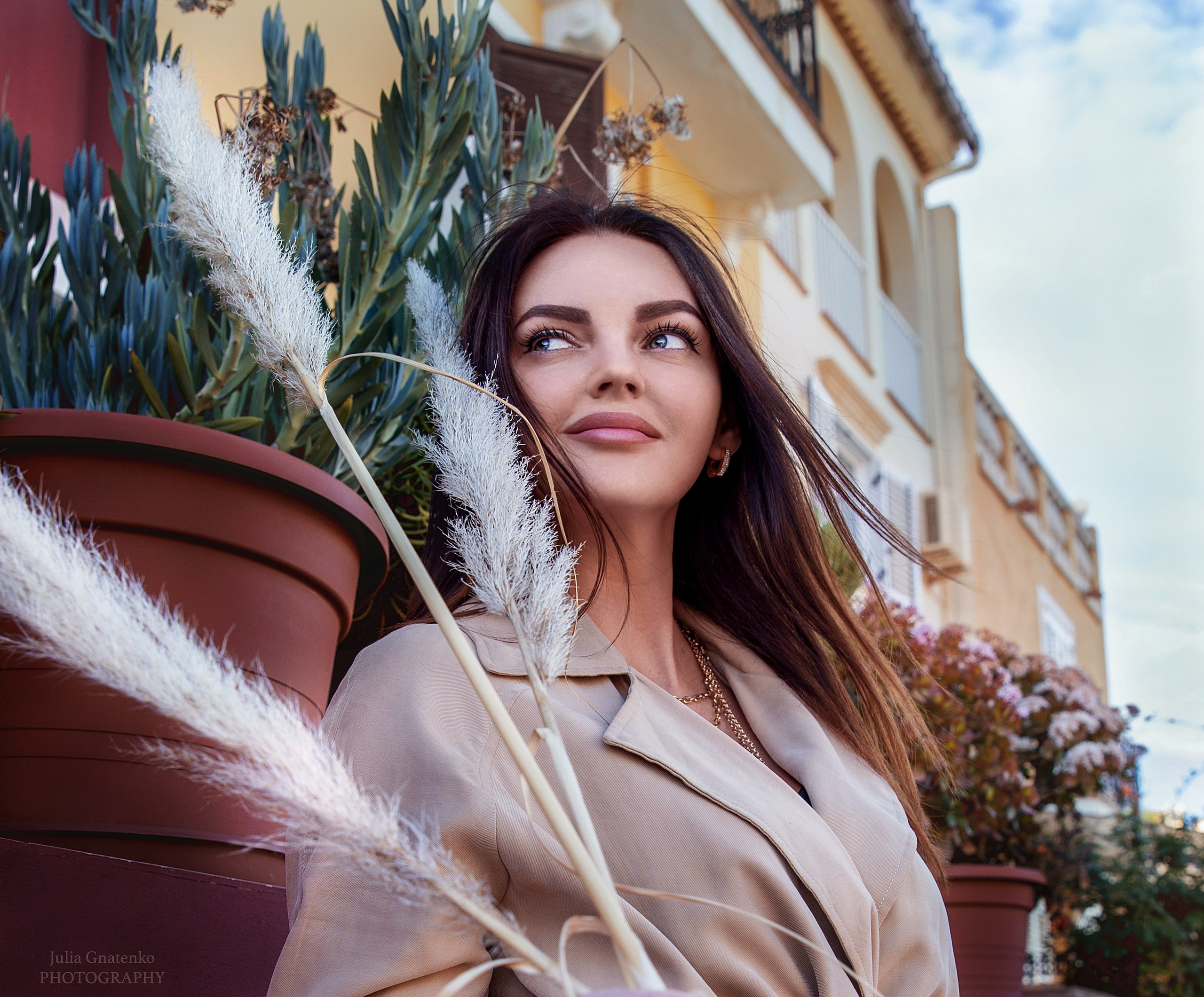 Модная женщина в бежевом пальто, держащая пампасную траву, позирует перед зданием в средиземноморском стиле с растениями и цветами в Валенсии, Испания
