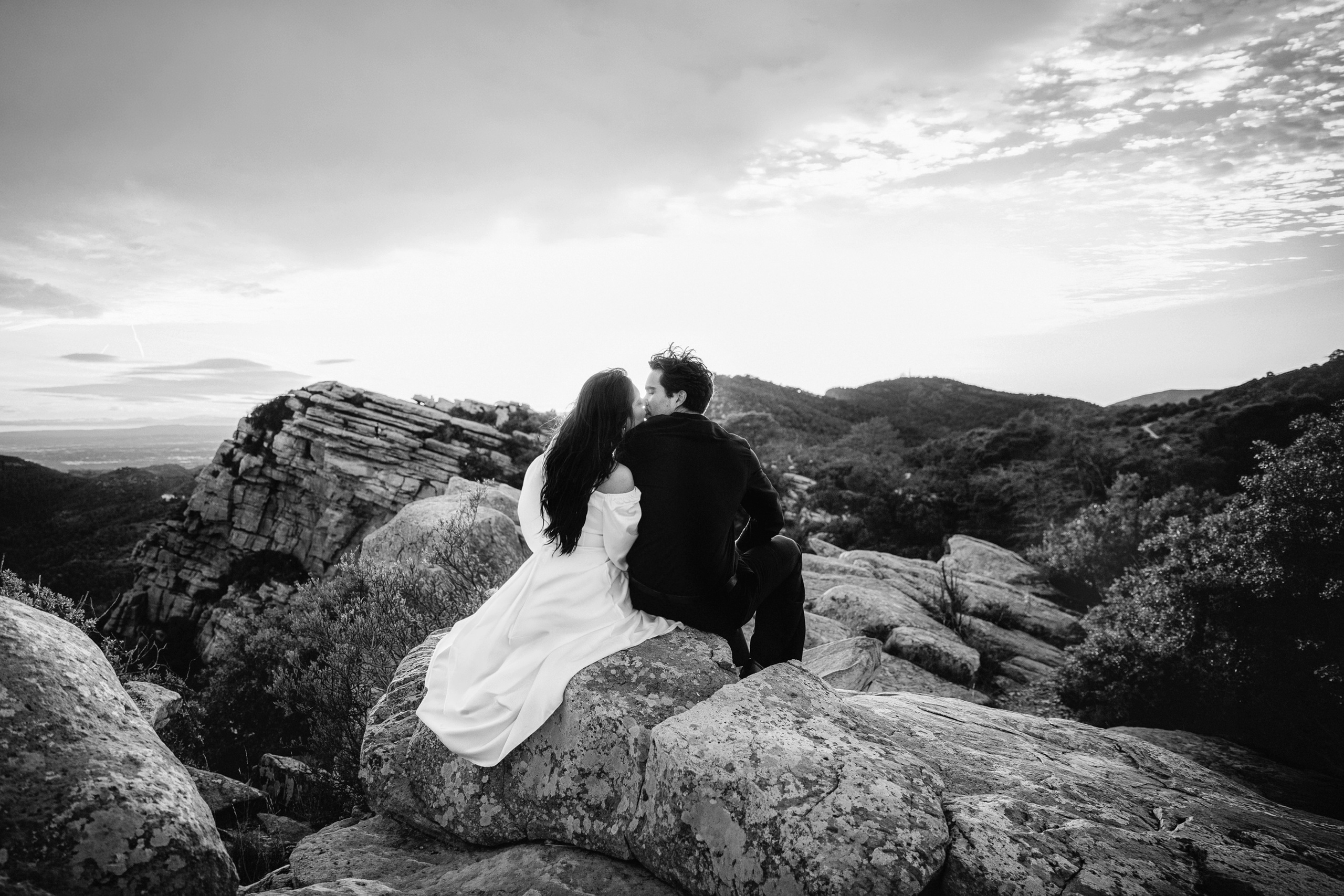 Bride and groom sitting together on natural stone formations during a secluded mountain destination wedding in Barcelona, Spain. This black and white elopement portrait highlights intimacy and timeless elegance.