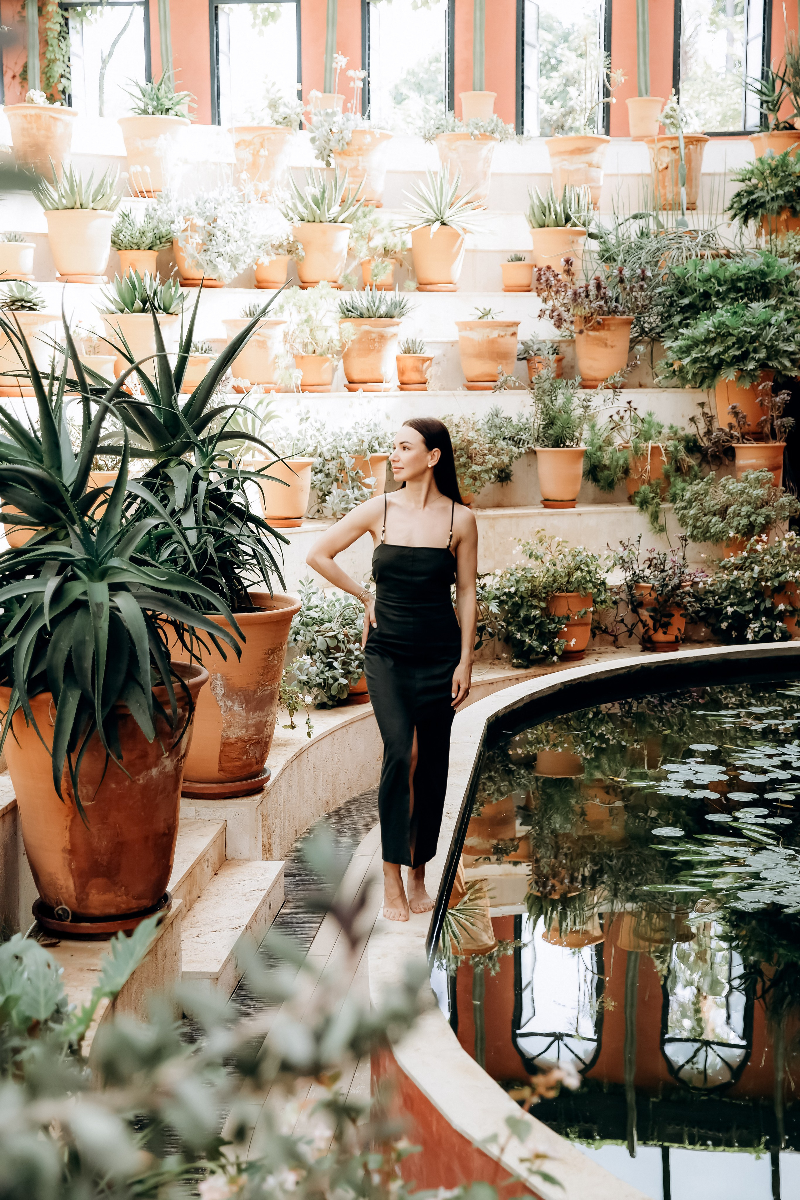 Женщина в черном платье стоит босиком у пруда с лилиями, окружённого ярусами терракотовых горшков с растениями в яркой средиземноморской оранжерее в Кальпе, Испания — ботанически вдохновленная портретная фотография, передающая природную красоту, спокойствие и художественную композицию, идеально подходит для портретных фотосессий в Кальпе и Испании.