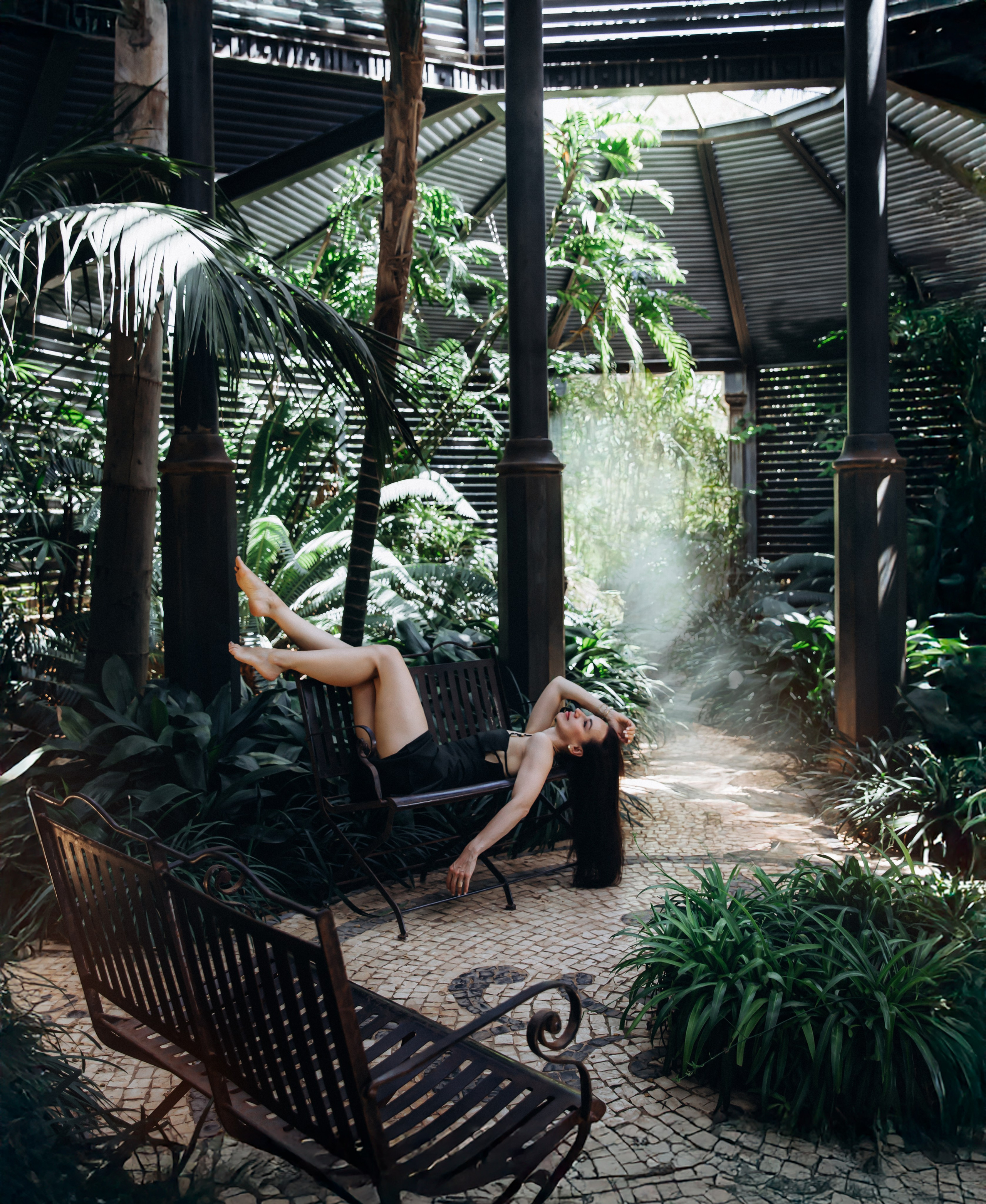 Женщина в черном платье лежит на кованой скамейке в пышном тропическом саду с драматичным светом и архитектурными тенями в Аликанте, Испания — креативная и выразительная портретная фотография, сочетающая элегантность, природу и современный дизайн, идеально подходит для портретных фотосессий в Аликанте и Испании.
