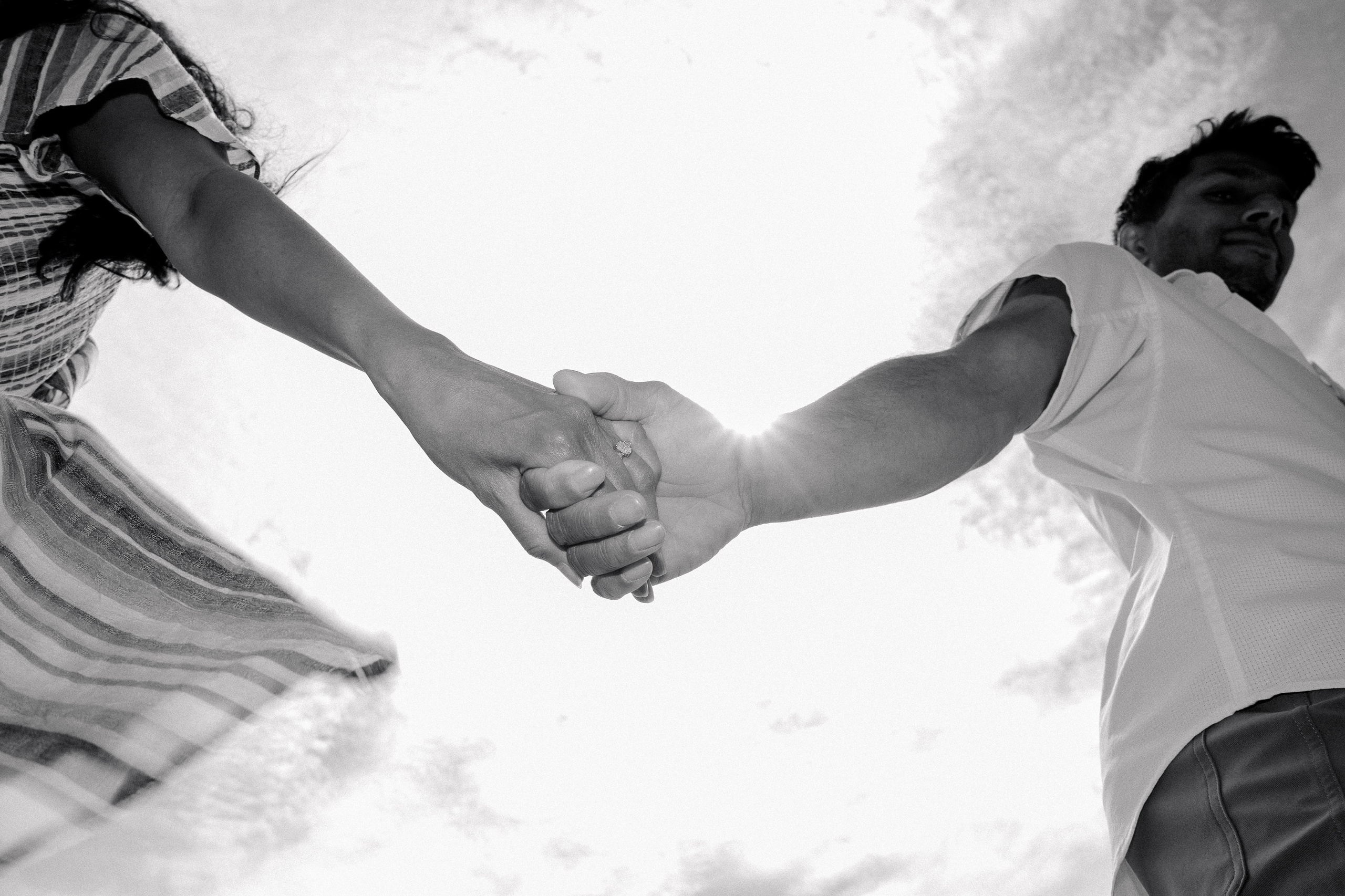 Dramatic low-angle black and white photo of an engaged couple holding hands under the bright sky, with sunlight highlighting the engagement ring — symbolizing connection and commitment.