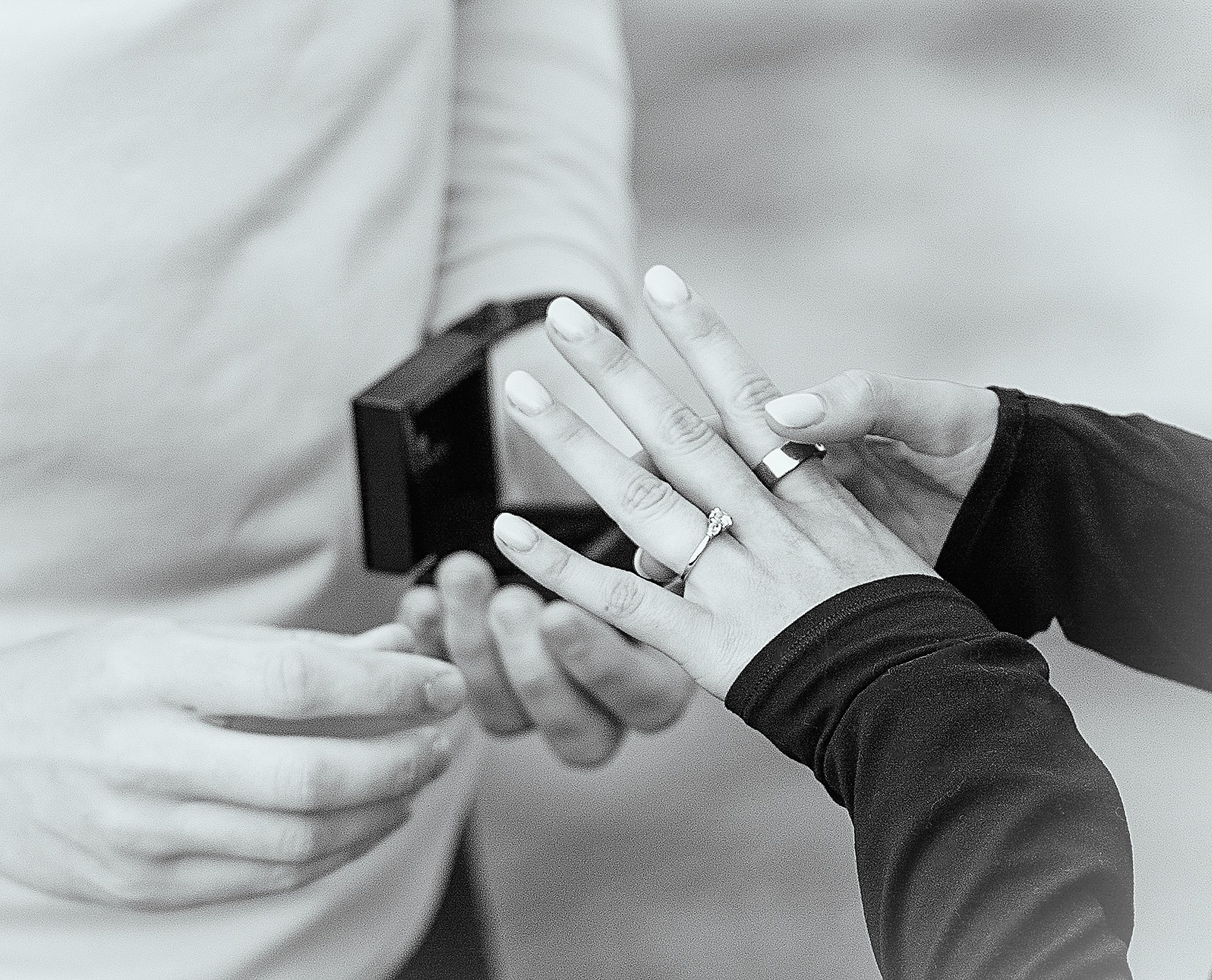 A close-up, black and white detail shot of a newly engaged couple holding hands to highlight a ring, set against an Irish landscape.
