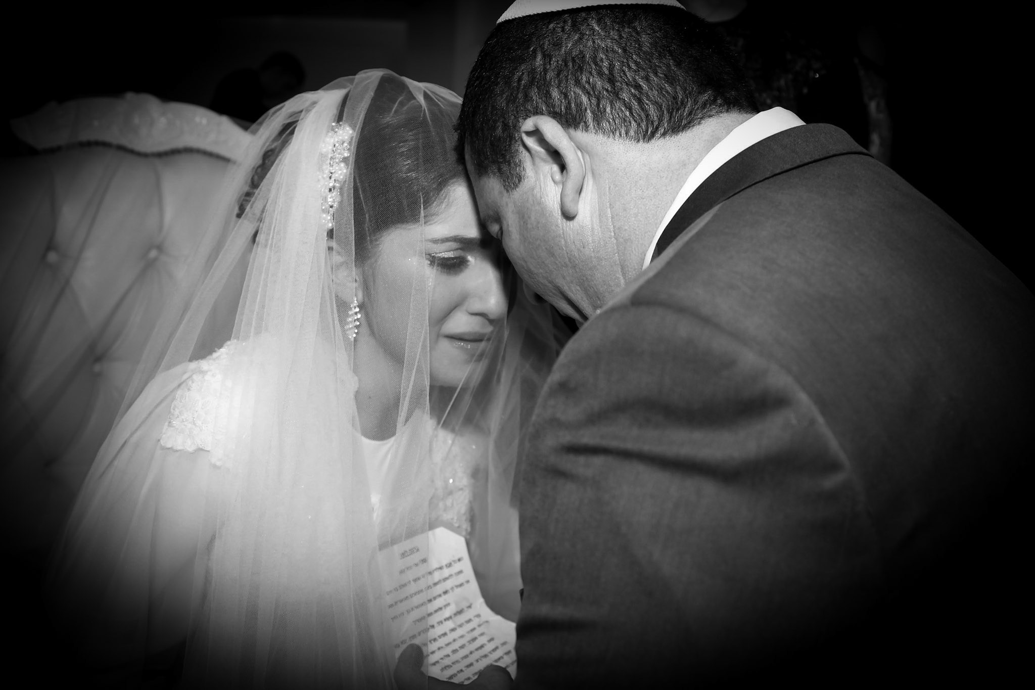 A tender moment at a wedding, with the bride in a white lace dress and veil, tearfully gazing as her father leans towards her. Their facial expressions and posture convey a deep emotional connection.