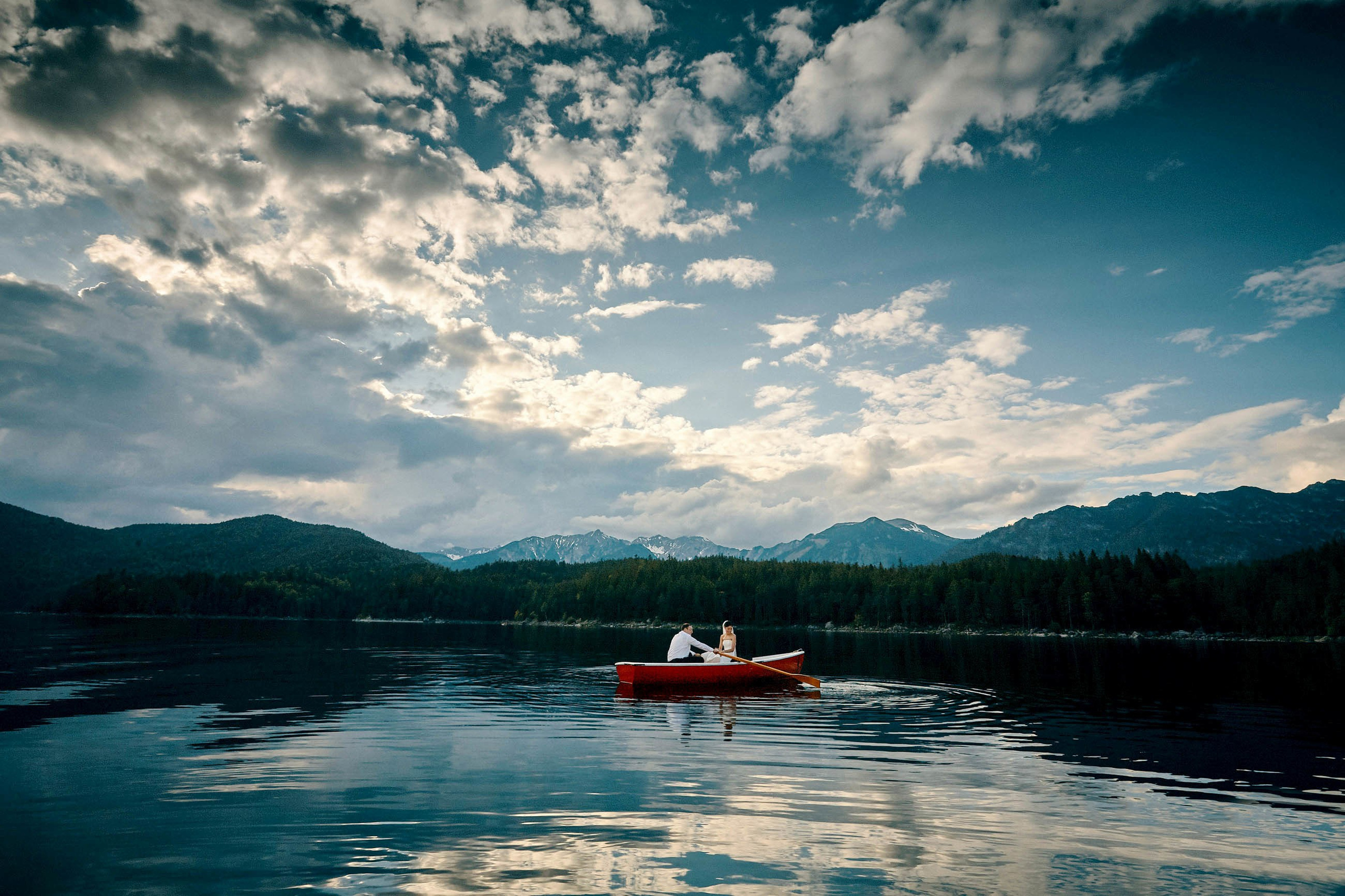 American couple in red rowboat on Lake Eibsee with Alps reflection, blue sky, and clouds.