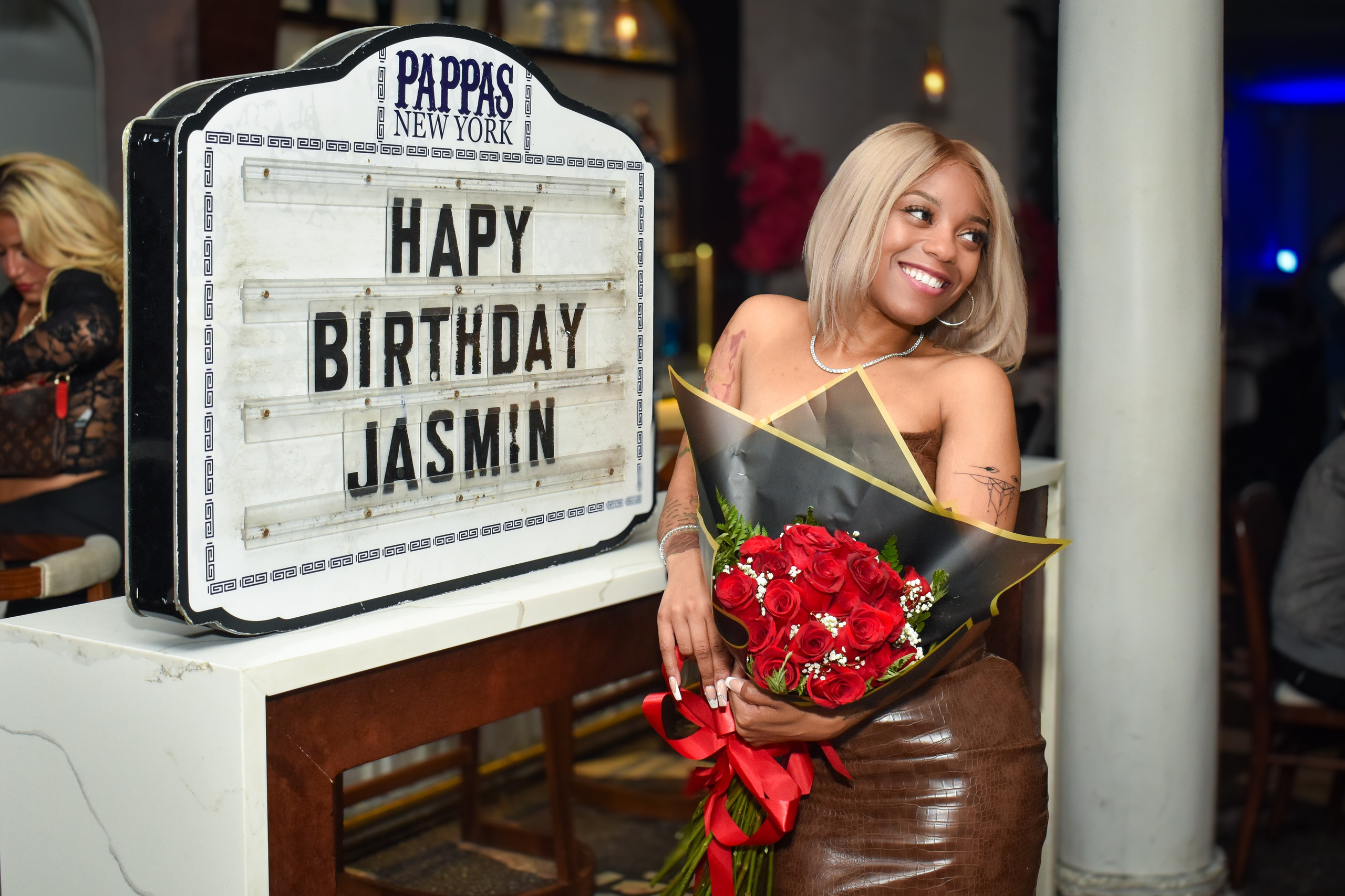 Woman holding a bouquet next to a “Happy Birthday” sign at a restaurant, NYC birthday photoshoot.