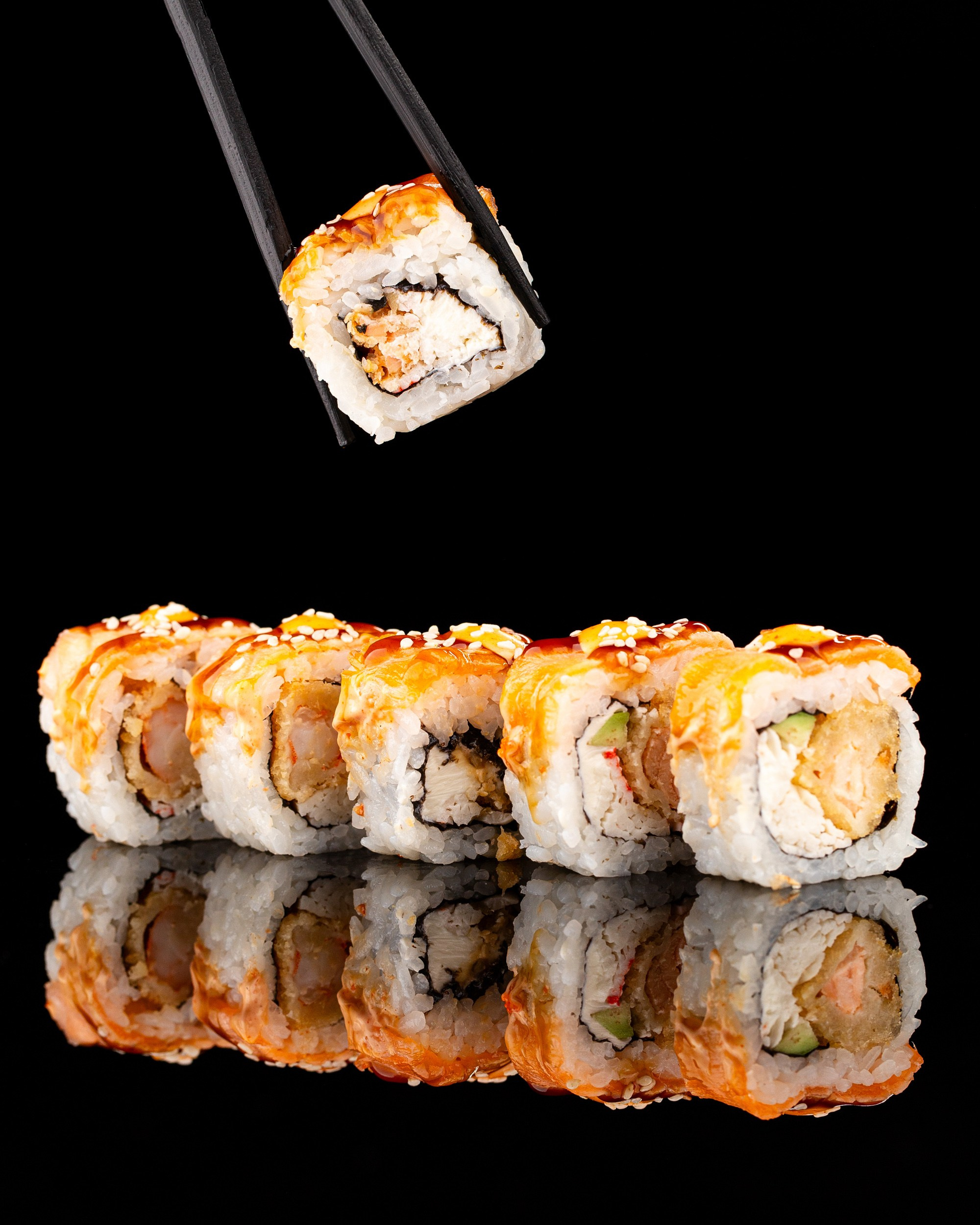 Qara fonda çubuqlarla tutulan suşi rulosu, küncüt toxumları və sousla bəzədilmiş