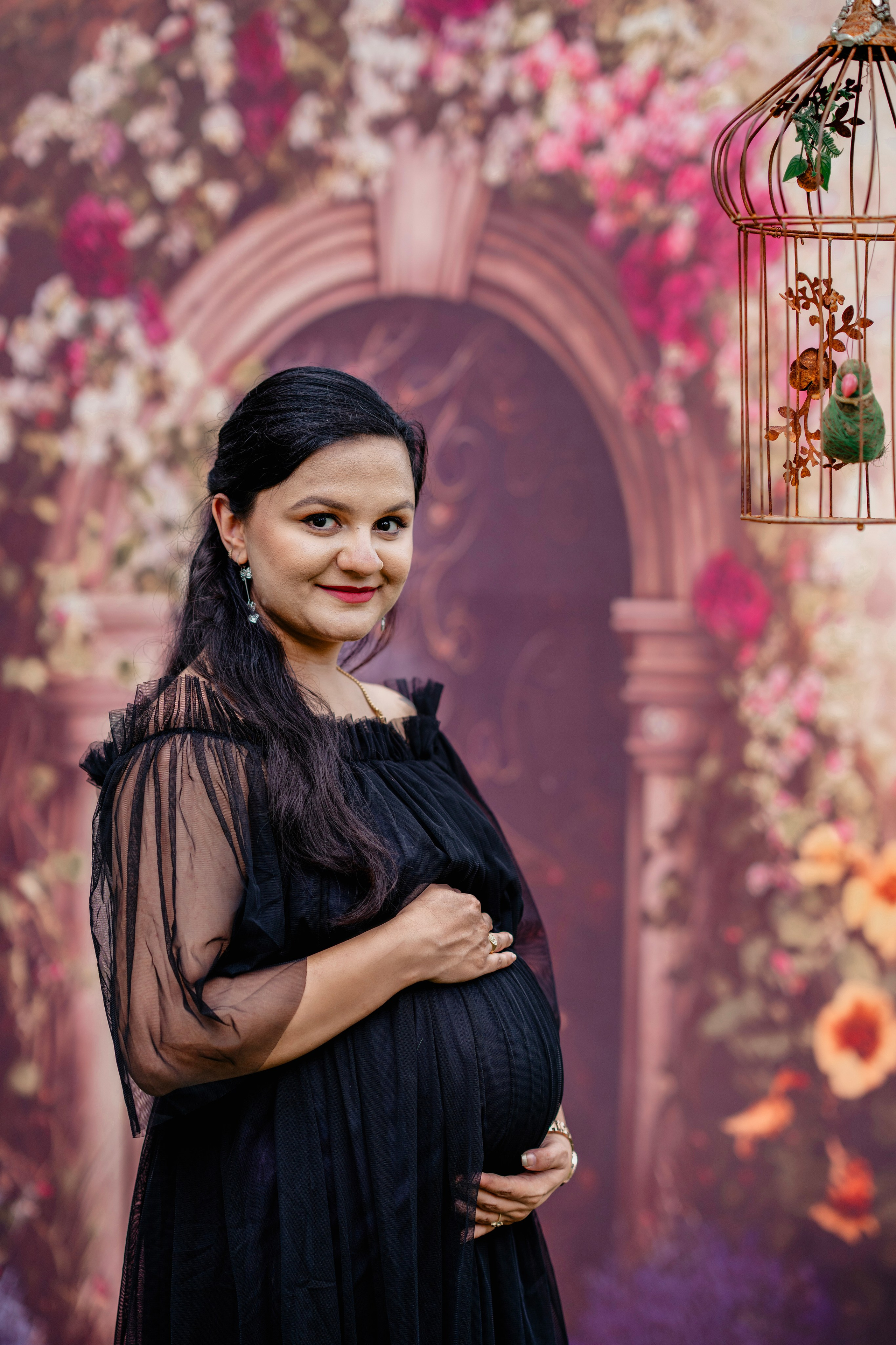 Maternity photoshoot in Bengaluru studio featuring a woman in a black off-the-shoulder tulle gown posing against a floral garden arch backdrop.