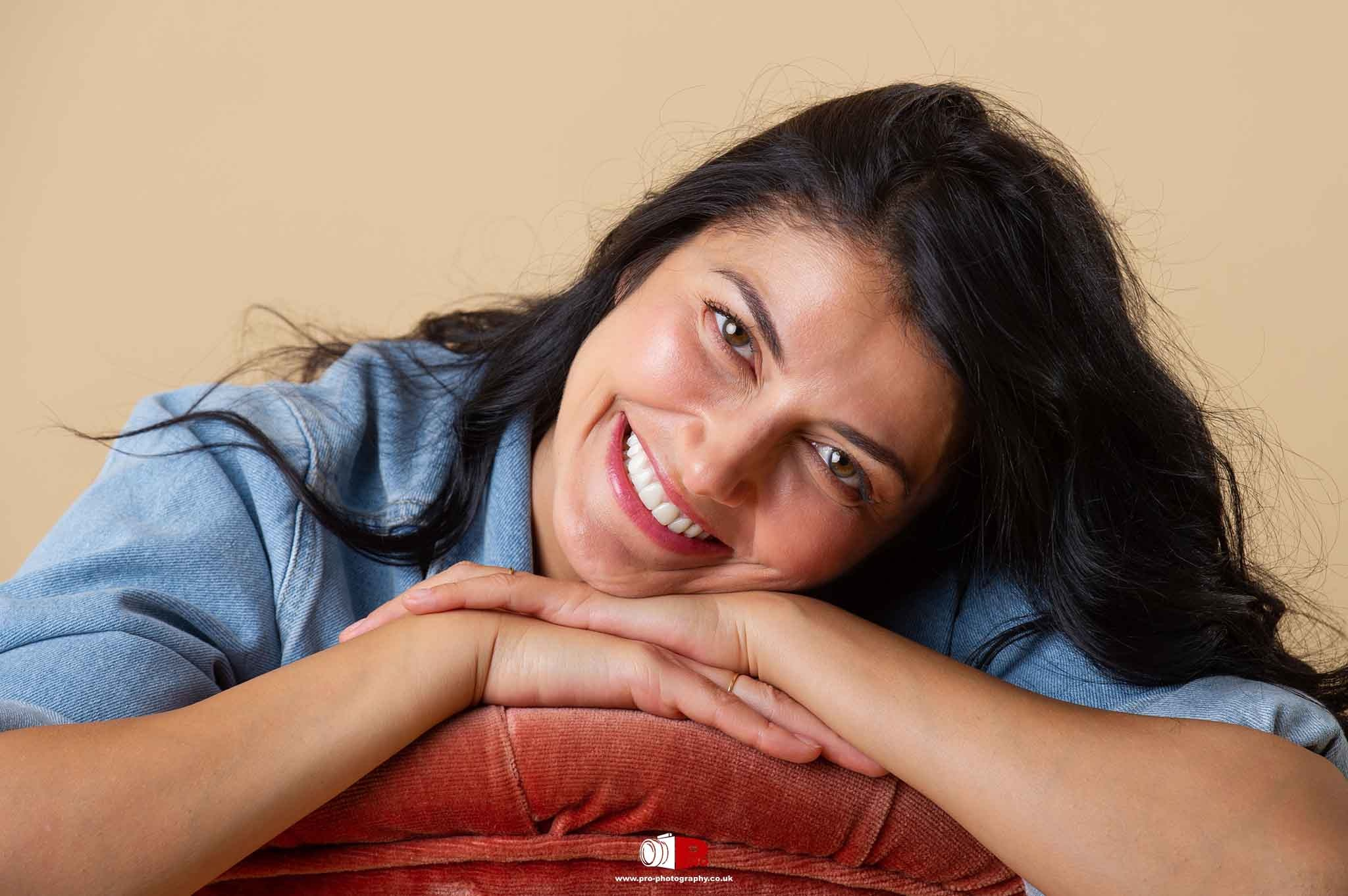 A happy woman in a casual denim shirt, resting her head on her hands over a rust-coloured cushion, smiling warmly.