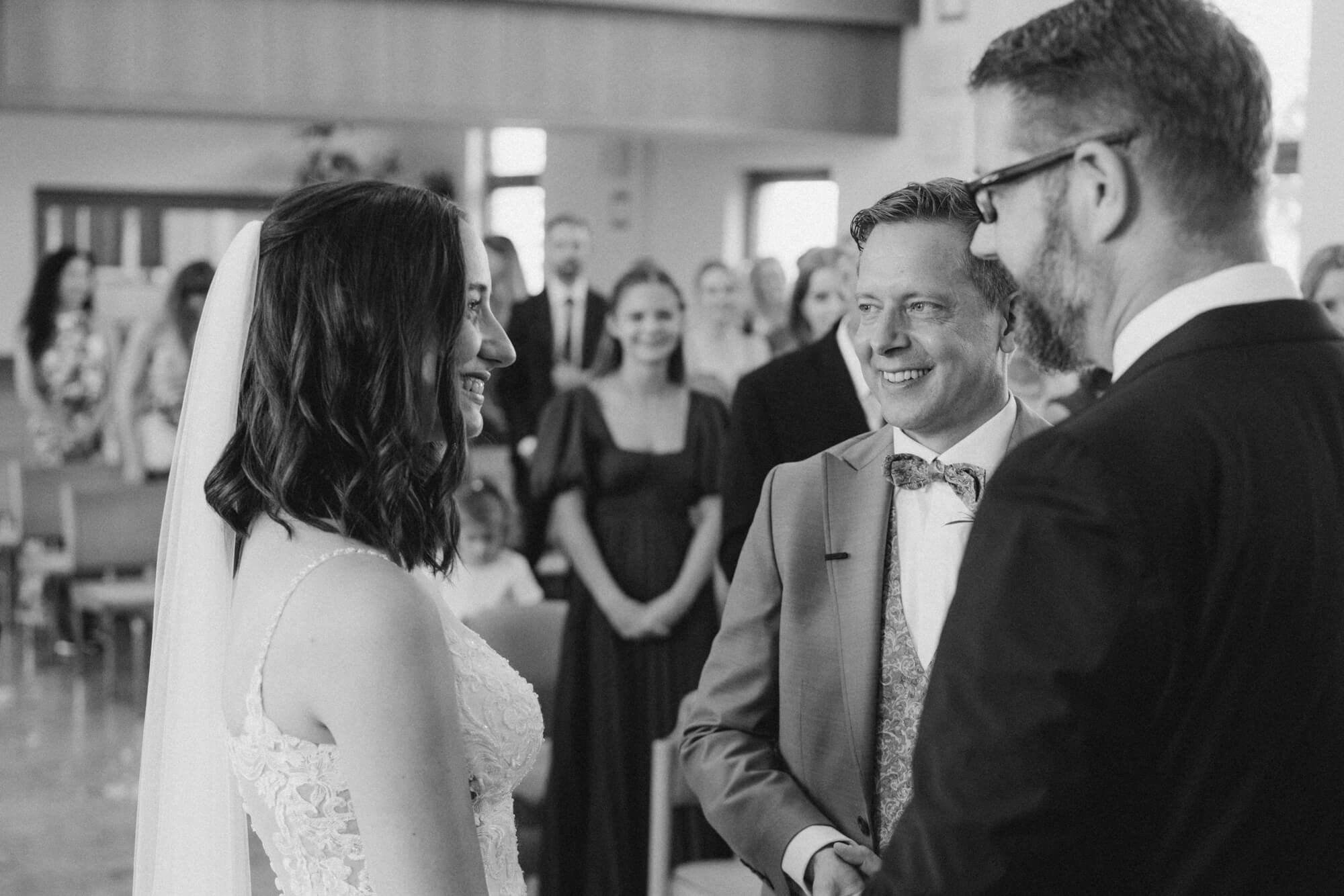 Bride and groom smiling at each other at the altar with a beaming guest in a bow tie visible beside them; black and white