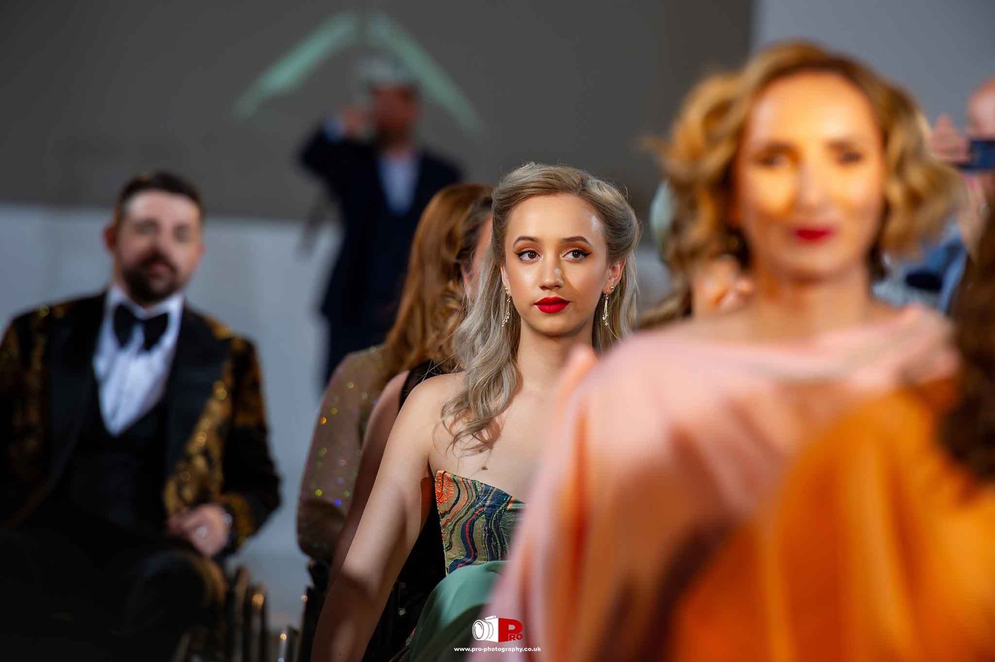 A young woman with platinum blonde hair attentively watching a fashion show with models in the background.
