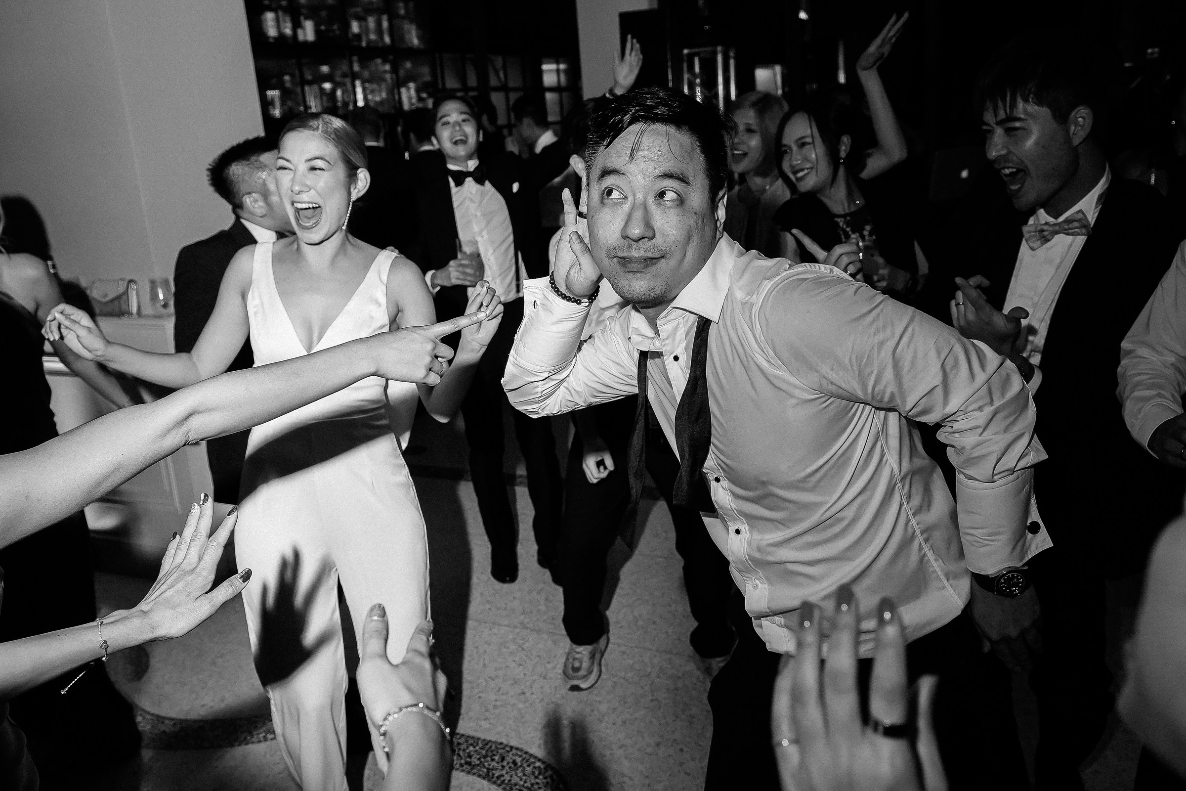 A lively wedding reception scene with a man playfully holding his ear as people, including the bride, laugh and point, creating a fun and joyous atmosphere.