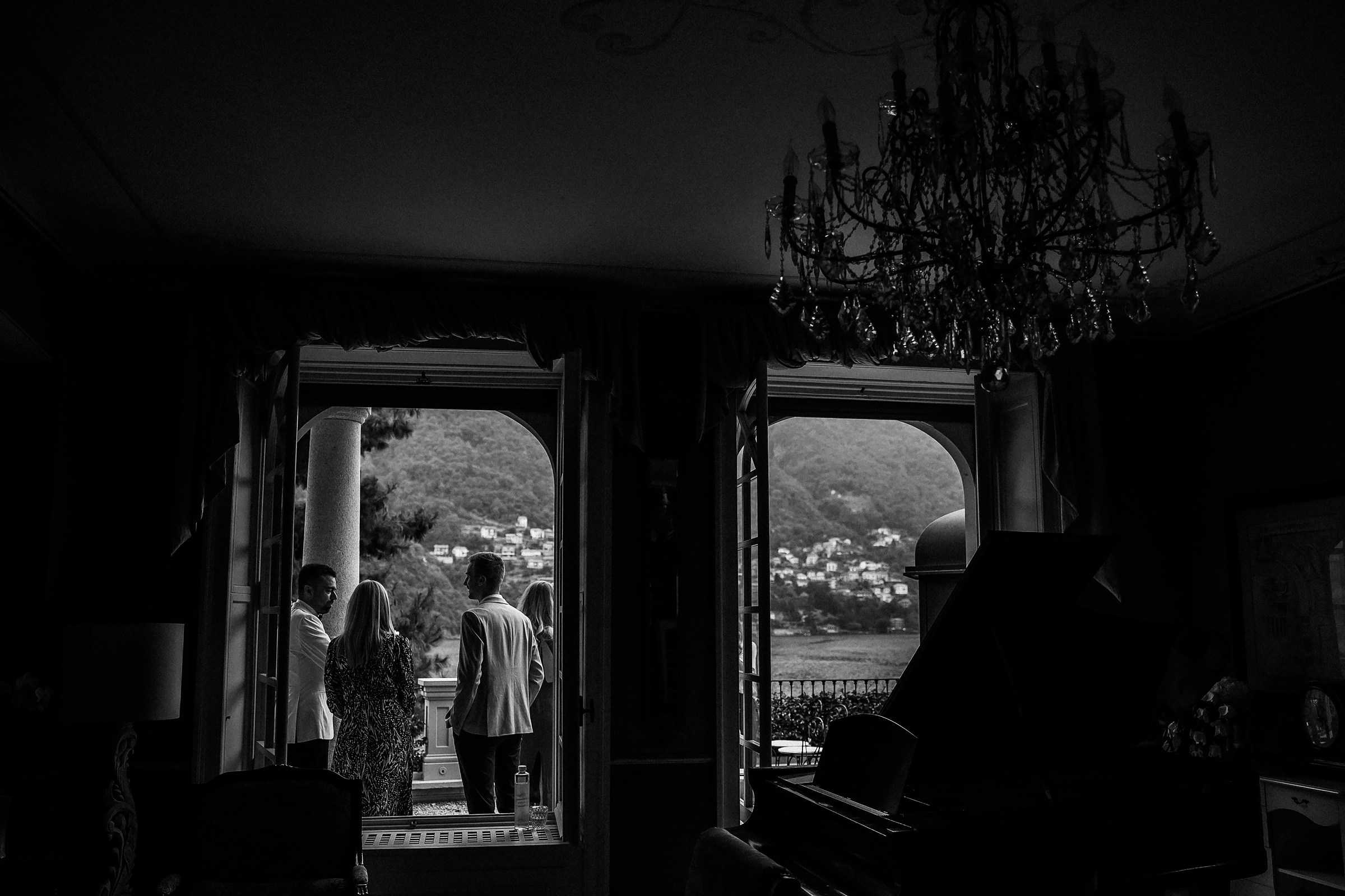 Black and white photo of wedding guests admiring the scenic view of Lake Como from a grand Relais Villa Vittoria interior with chandelier and piano.
