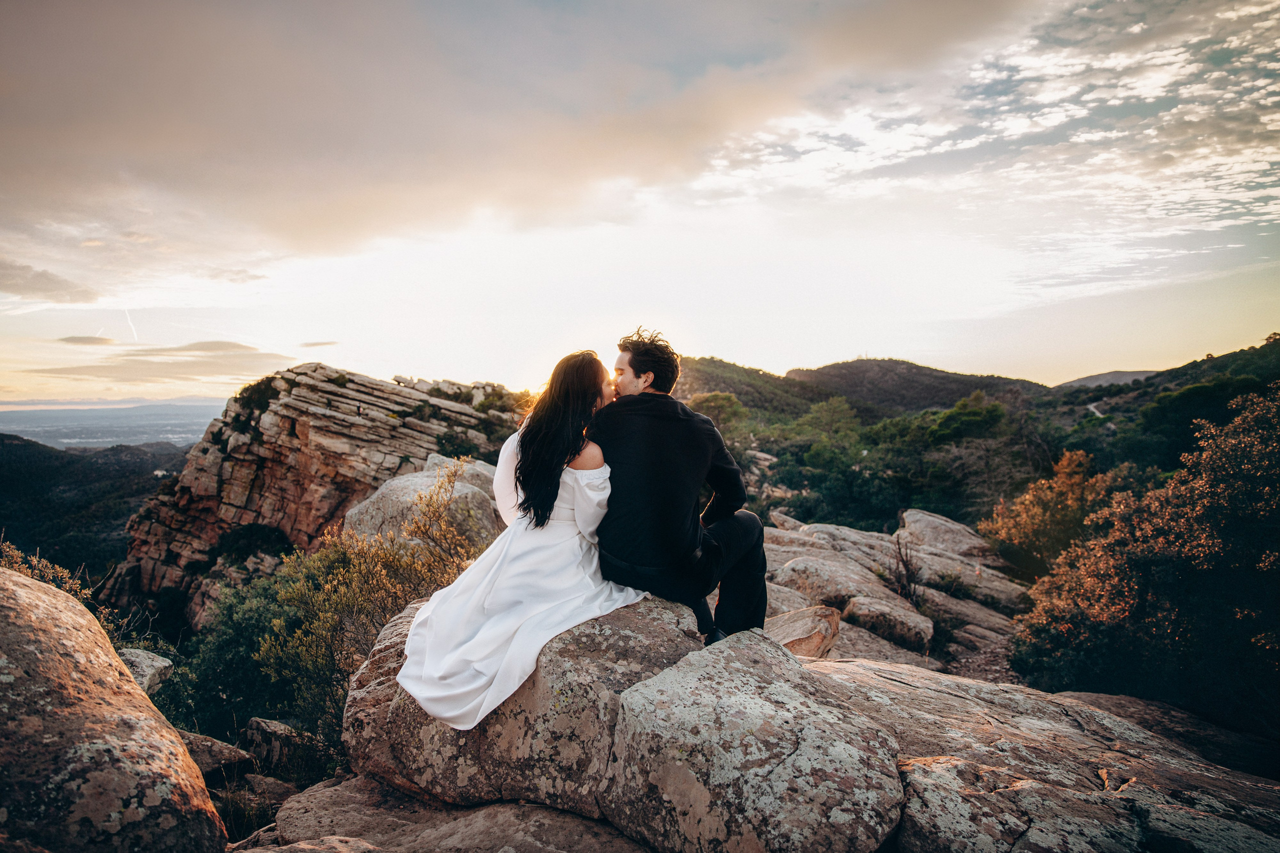 Newlyweds sitting together on natural rock formations during a secluded mountain elopement in Barcelona, Spain. The wide landscape and cloudy sky emphasize the intimacy of their destination wedding.