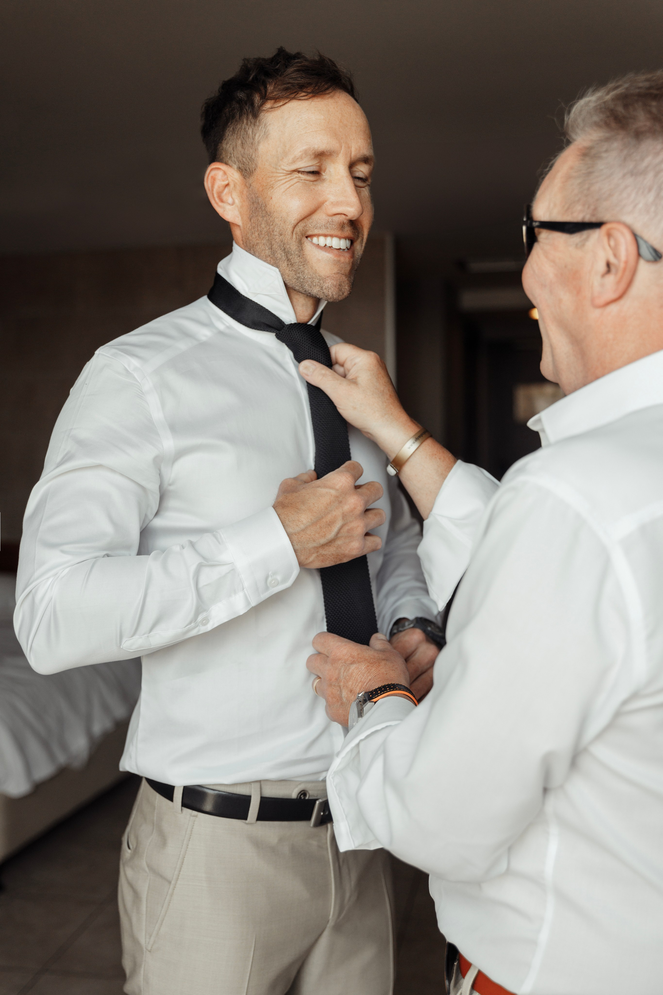 The groom getting ready in a cozy room, with his father helping him put on his tie, on the big day at the hotel in Rhodes, Greece