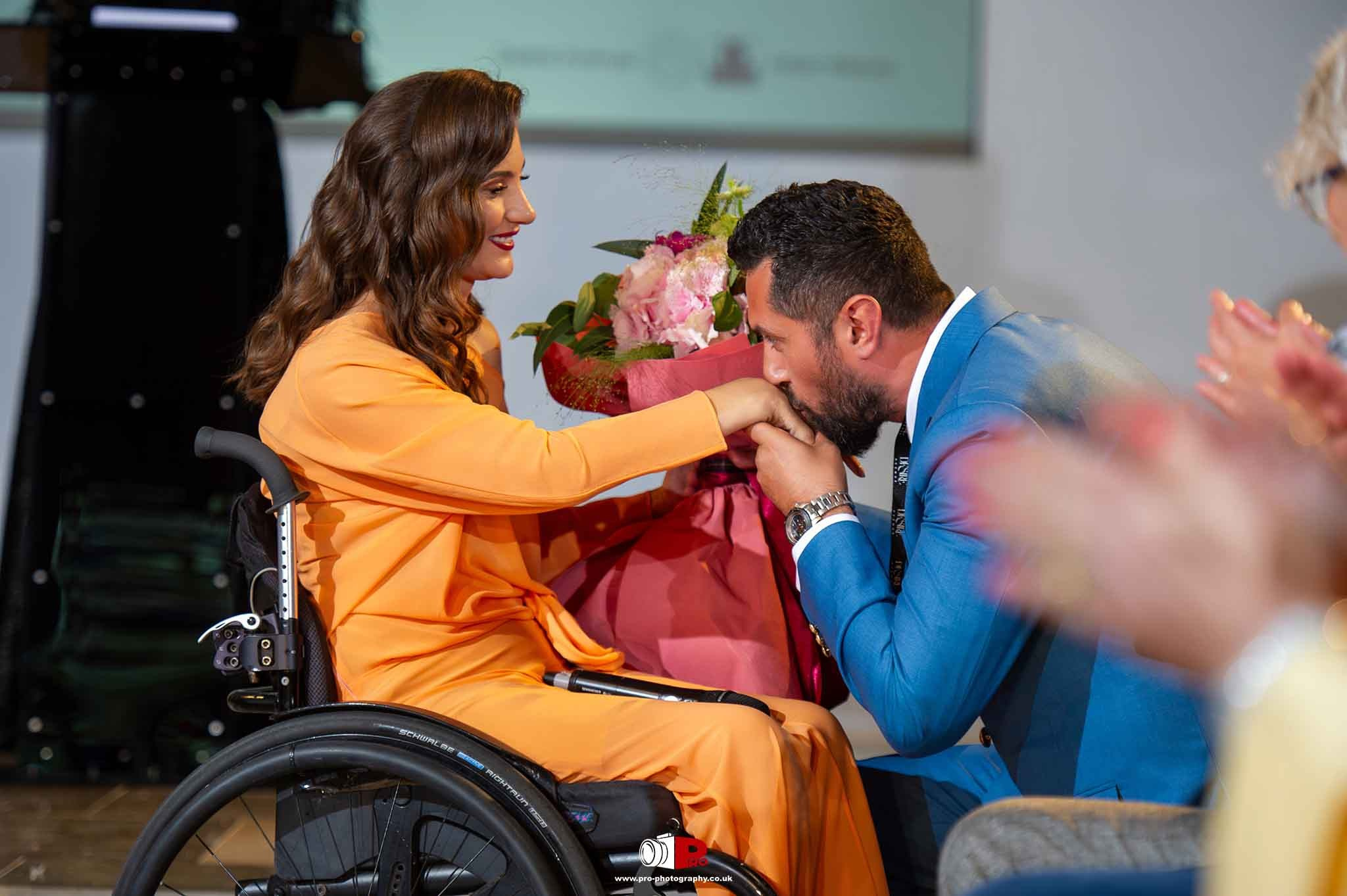A man in a blue suit kisses a woman's hand in an orange dress seated in a wheelchair, surrounded by applauding guests.