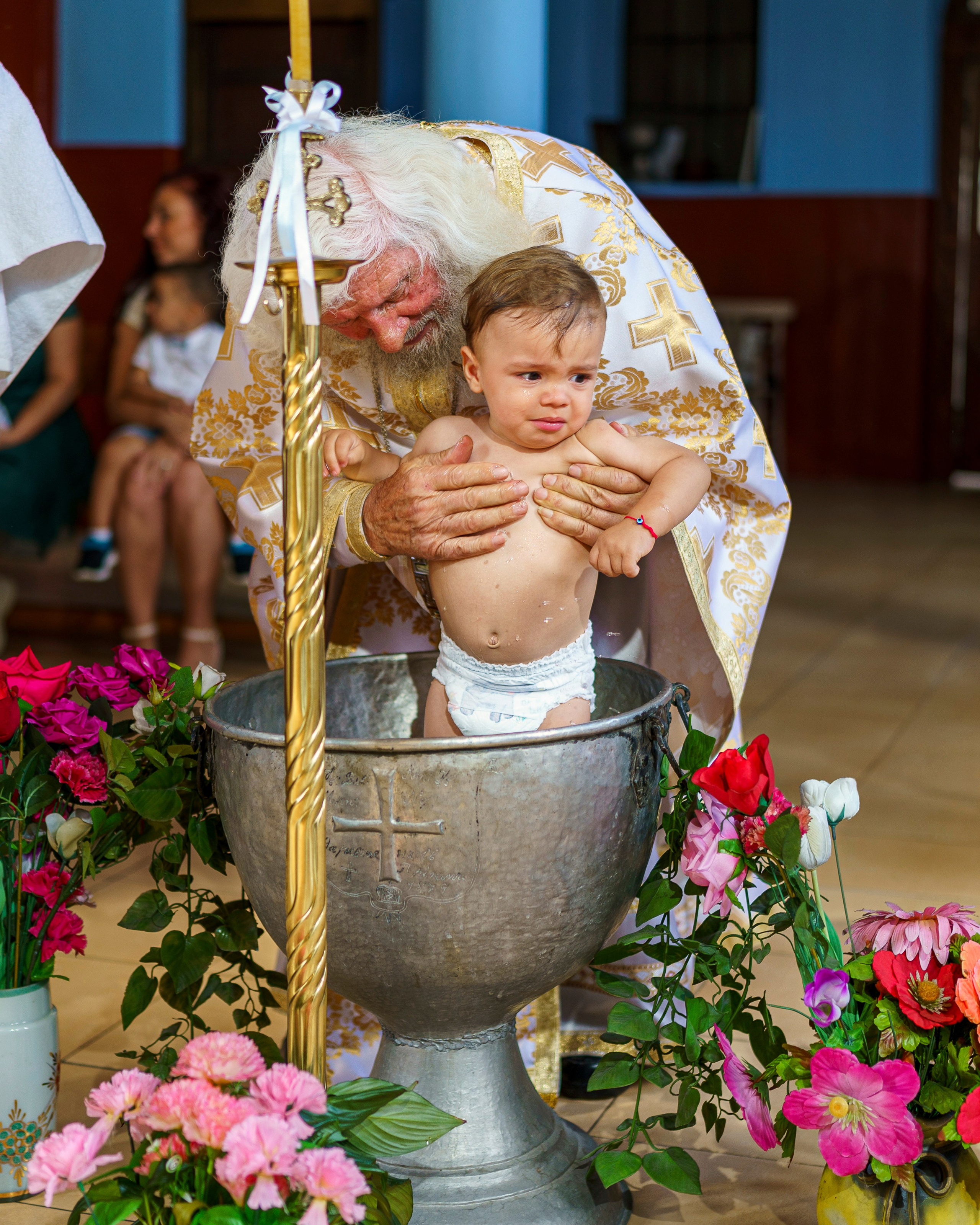 Православно кръщене на бебе в църква – свещеник извършва тайнството над купел, украсен с цветя.