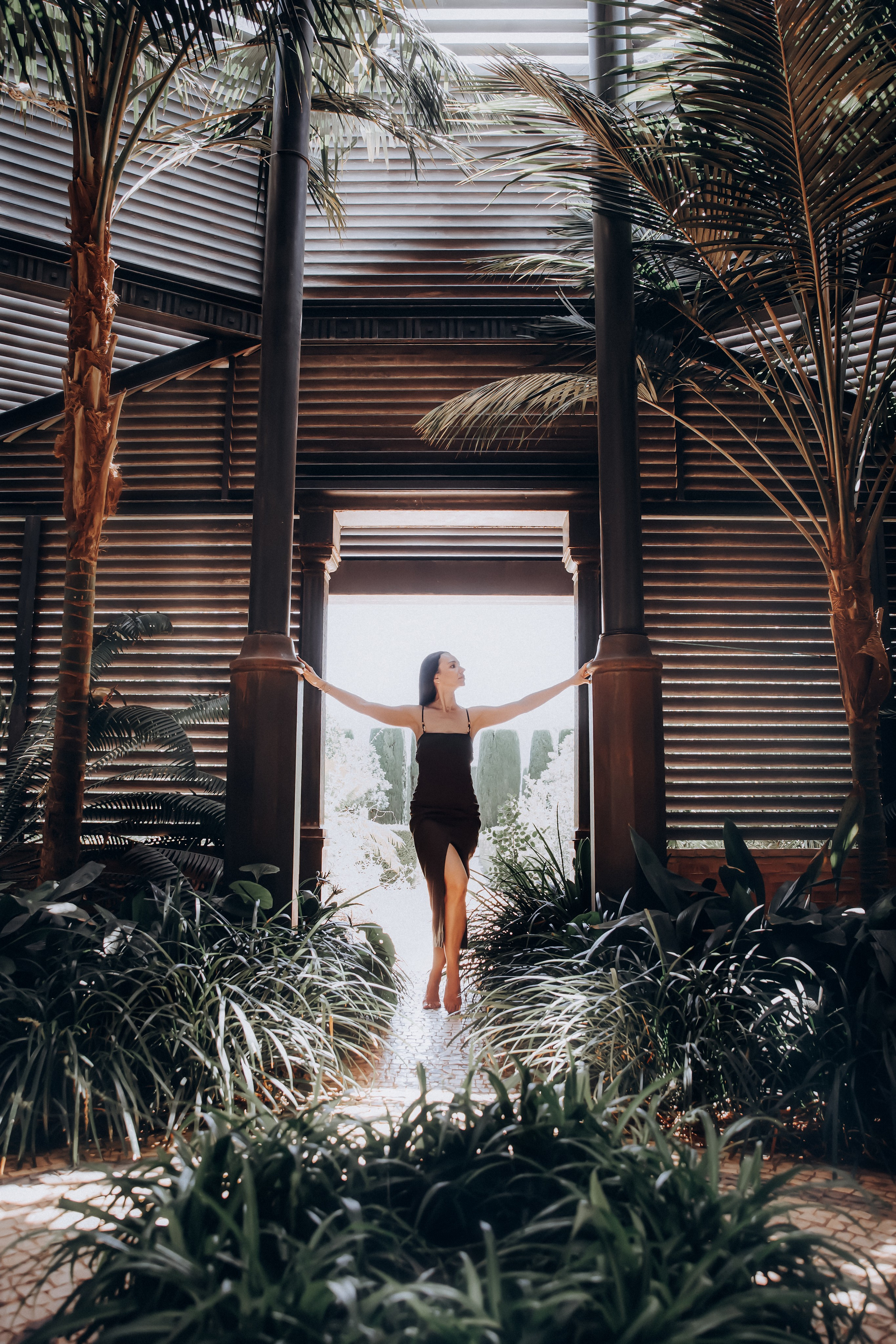 Элегантная женщина в черном платье уверенно позирует в тропическом зимнем саду с архитектурными элементами в Барселоне, Испания — современная портретная фотография, передающая естественный свет, модный стиль и спокойную атмосферу.