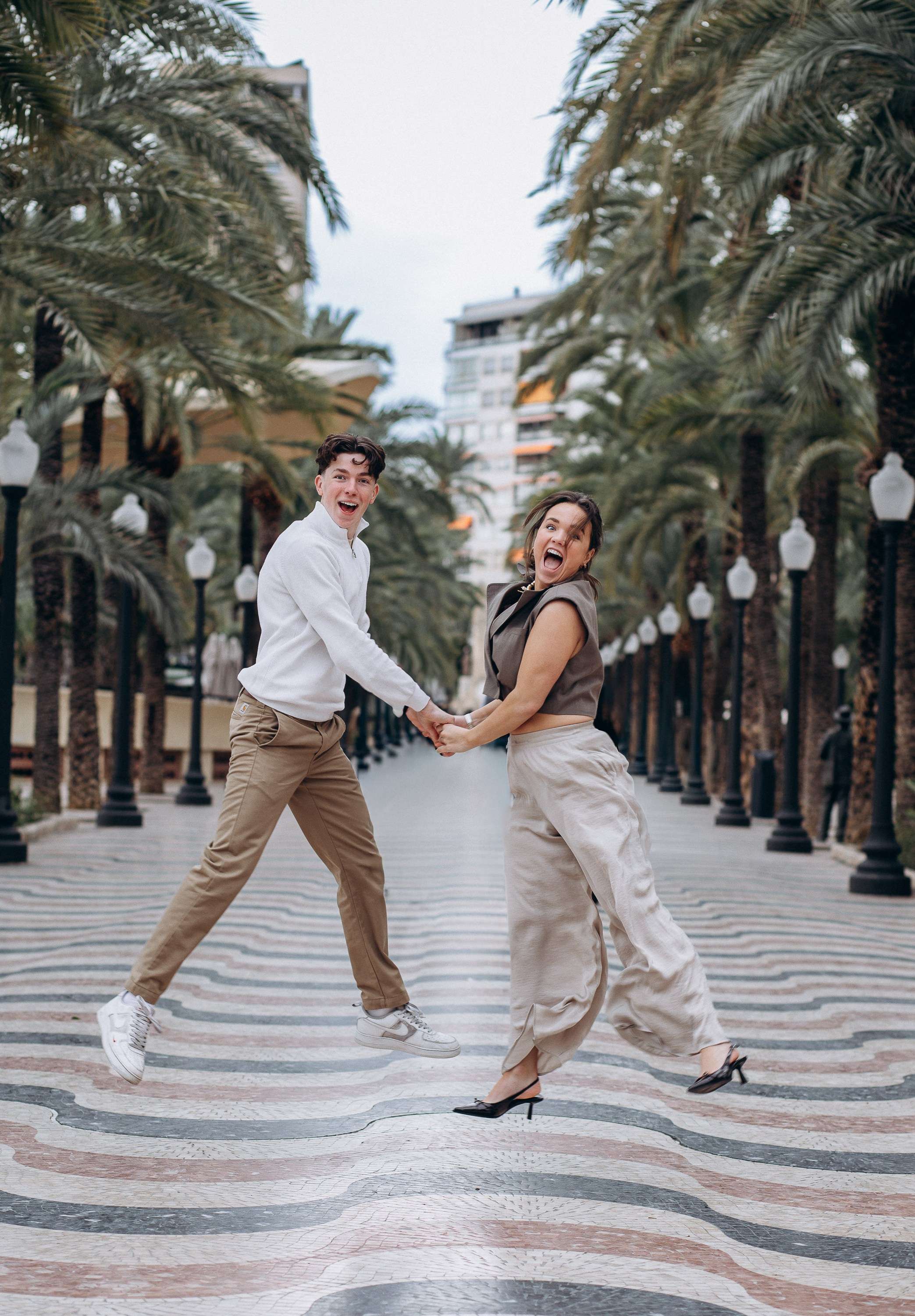 Sesión de fotos familiar en Alicante, España, que captura un momento alegre y dinámico de una madre y su hijo adolescente caminando de la mano y riendo juntos en un paseo rodeado de palmeras, fotografiado con luz natural y arquitectura mediterránea, creando una atmósfera enérgica y divertida — ideal para fotografía familiar, sesiones familiares lifestyle, retratos familiares espontáneos y sesiones familiares emotivas en Alicante y en toda España.