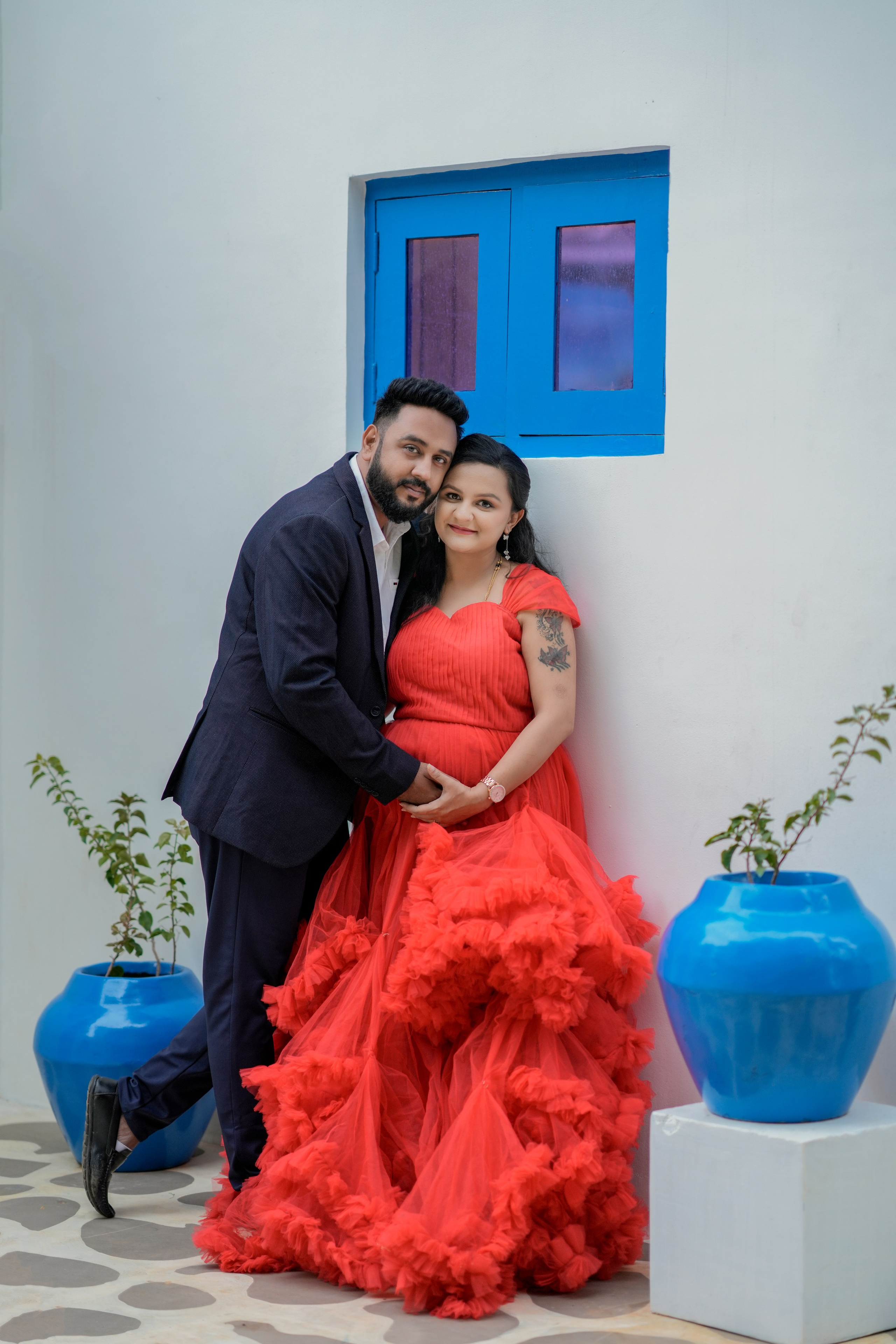 Maternity photoshoot in Bengaluru featuring a man in a navy suit and a woman in a red ruffled gown posing by a blue window and blue clay pots.
