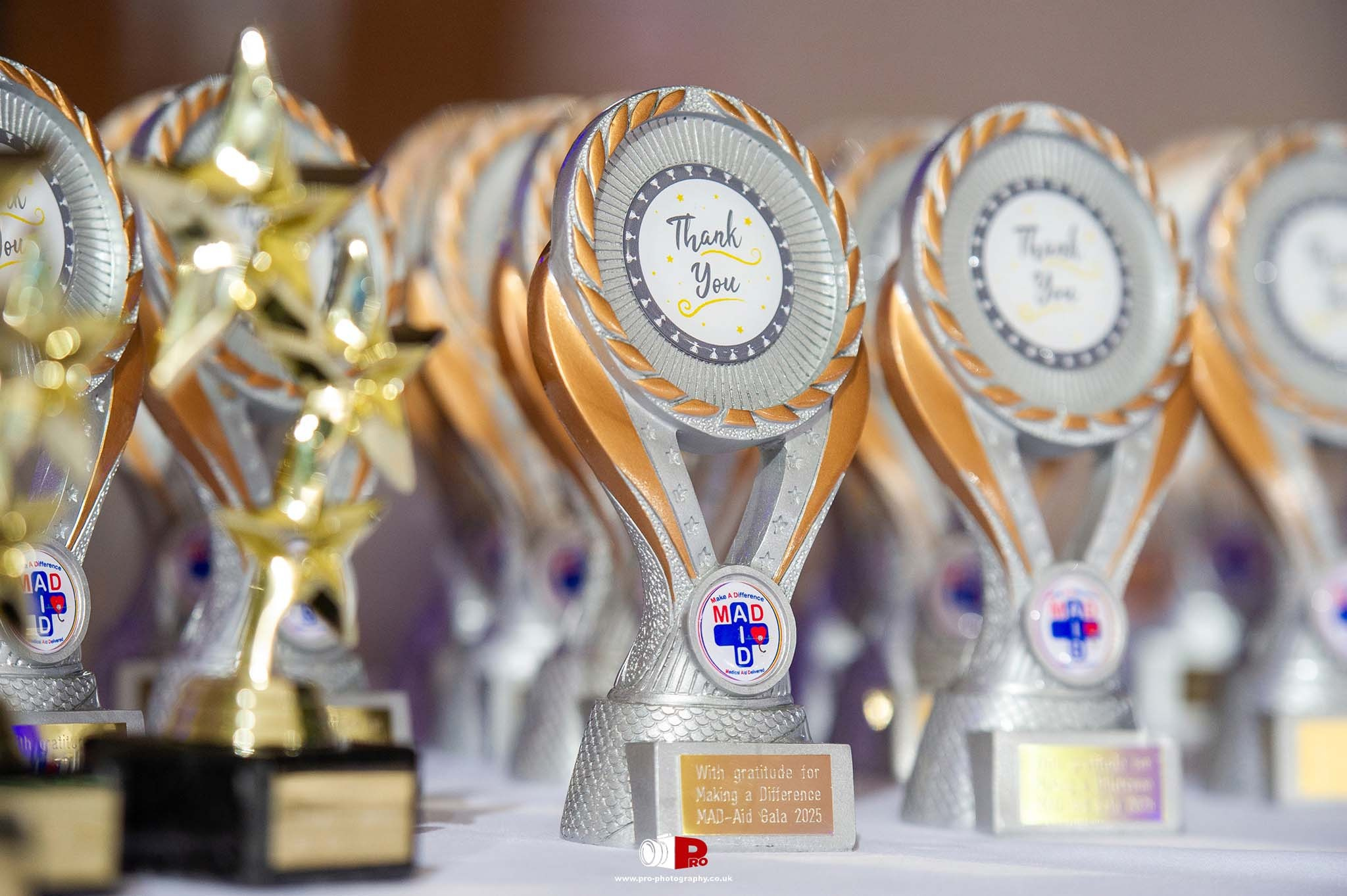 Close-up of MAD-Aid Gala 2025 "Thank You" trophies with gold and silver details displayed on a table.