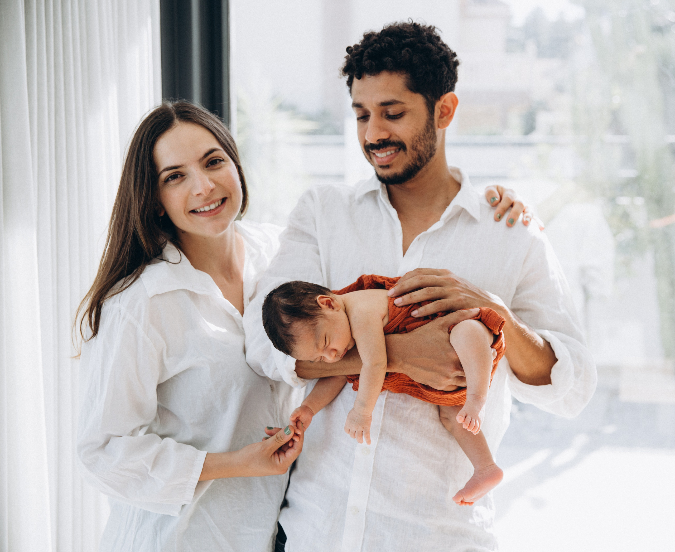 Retrato familiar alegre en interiores en Alicante, España — pareja sonriente vestida de blanco sosteniendo a su bebé recién nacido bajo una luz natural suave. Perfecto para familias que buscan sesiones lifestyle en Alicante y España que capturen calidez y ternura.