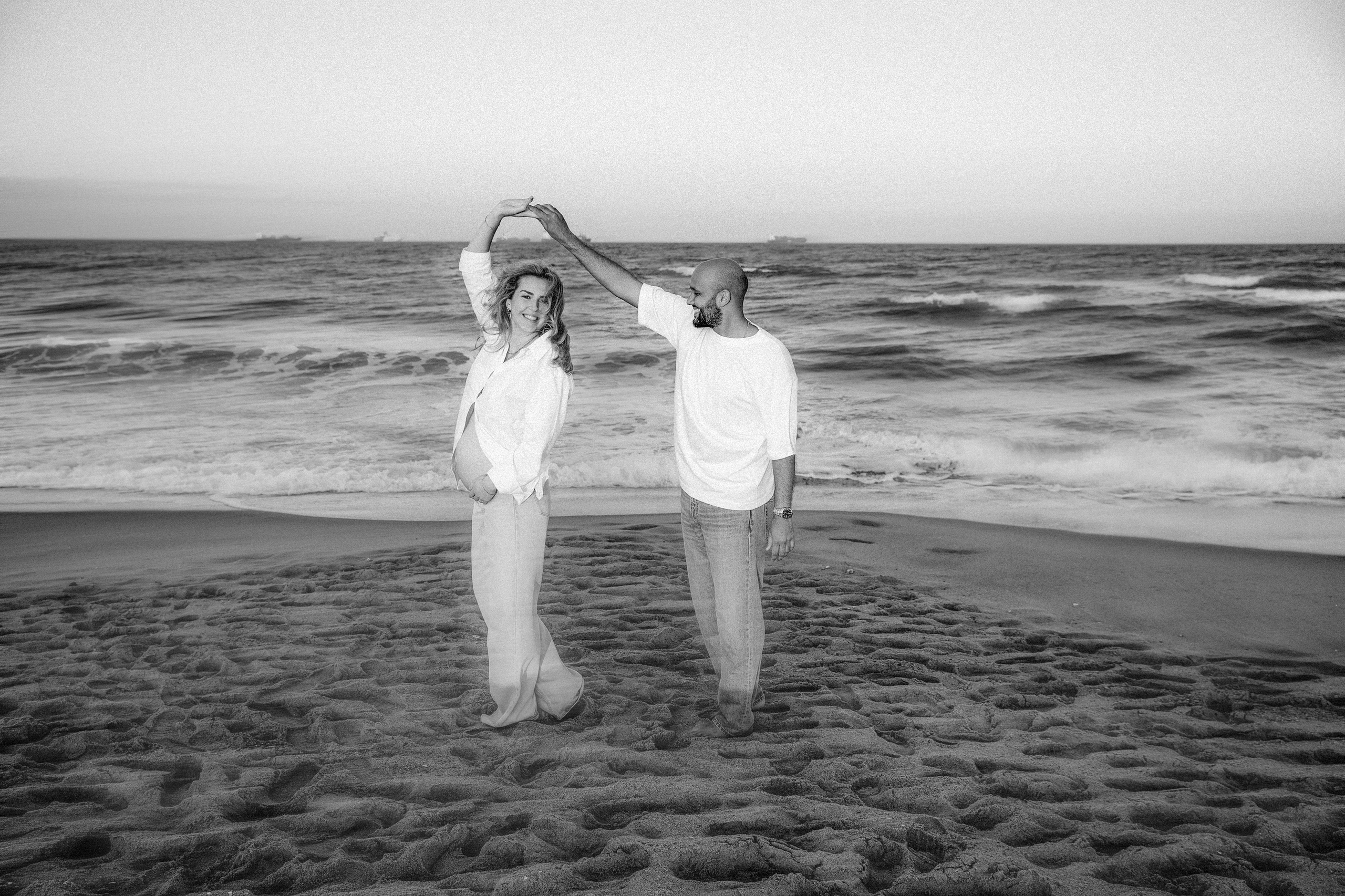 Sesión de fotos de embarazo en Valencia, España, con una pareja cogida de la mano en la playa en blanco y negro, creando un momento de embarazo emotivo y atemporal junto al mar — ideal para sesiones maternity y de pareja en Valencia y en toda España.