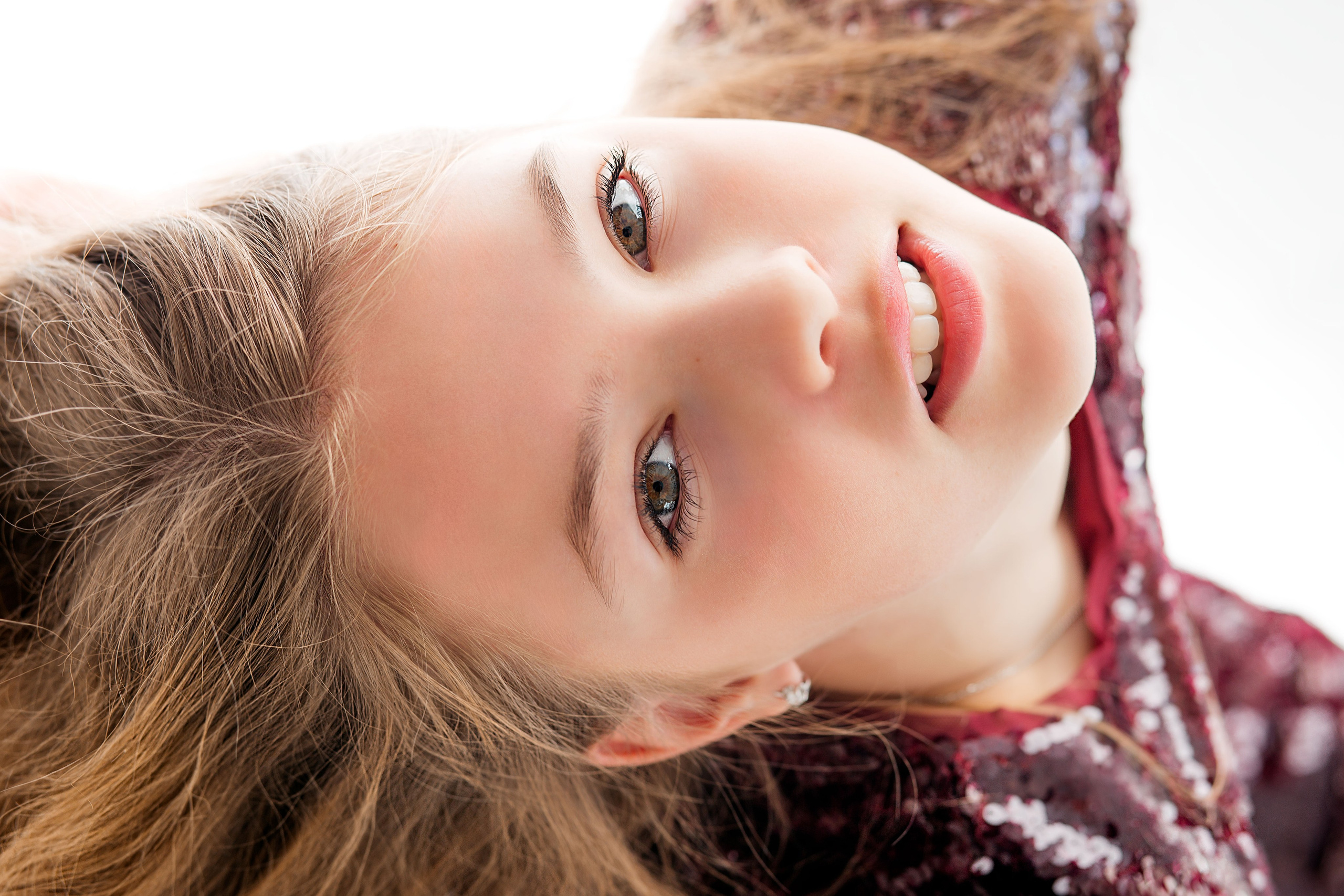 Крупный портрет улыбающейся девочки в блестящем бордовом платье, лежащей и смотрящей в камеру с яркими глазами и распущенными волосами, в студии в Валенсии, Испания