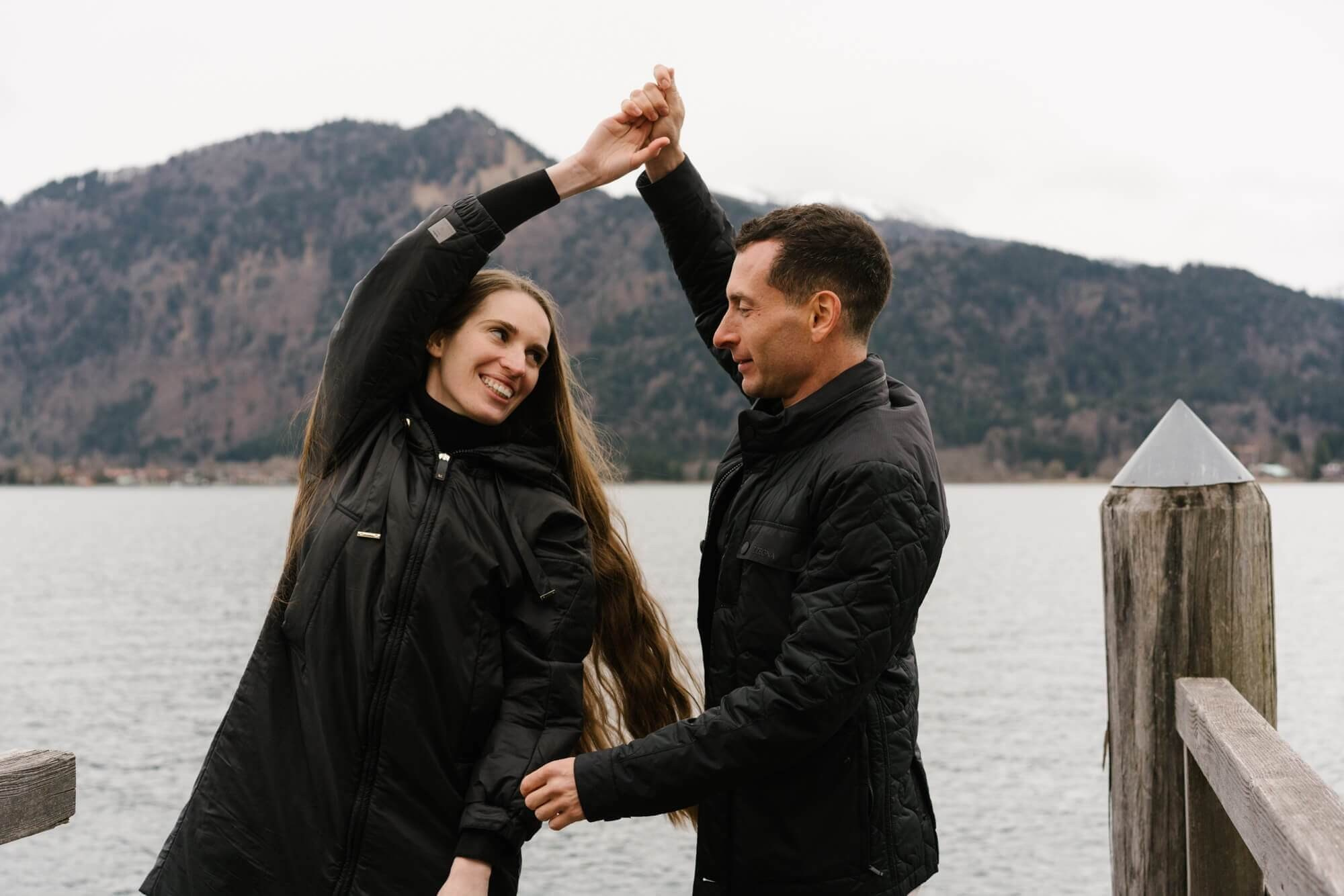 Man spinning and dancing with woman on a wooden dock at Tegernsee lake during playful couple photo session Germany