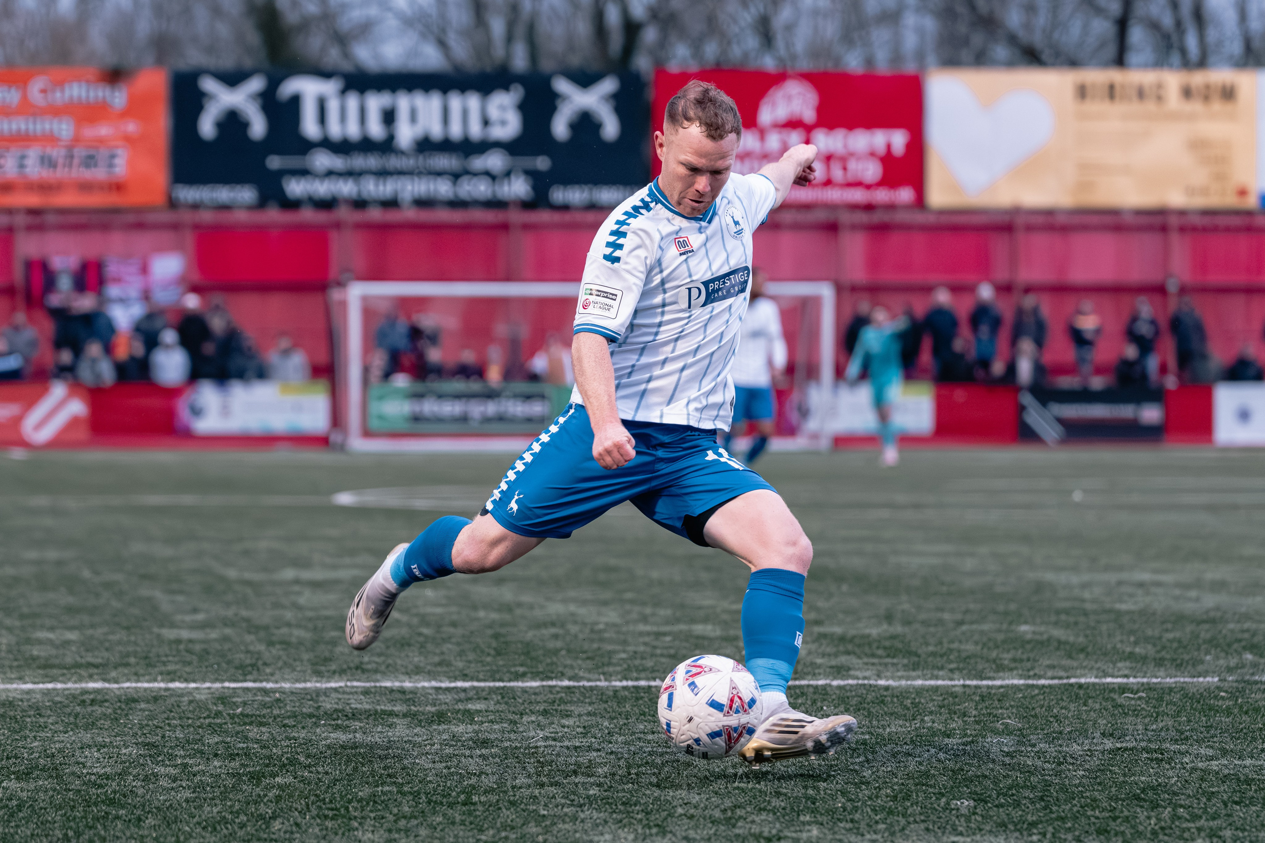 Hartlepool United F.C. forward Adam Campbell swings his right foot through the ball to shoot, with the red stand and spectators in the background.