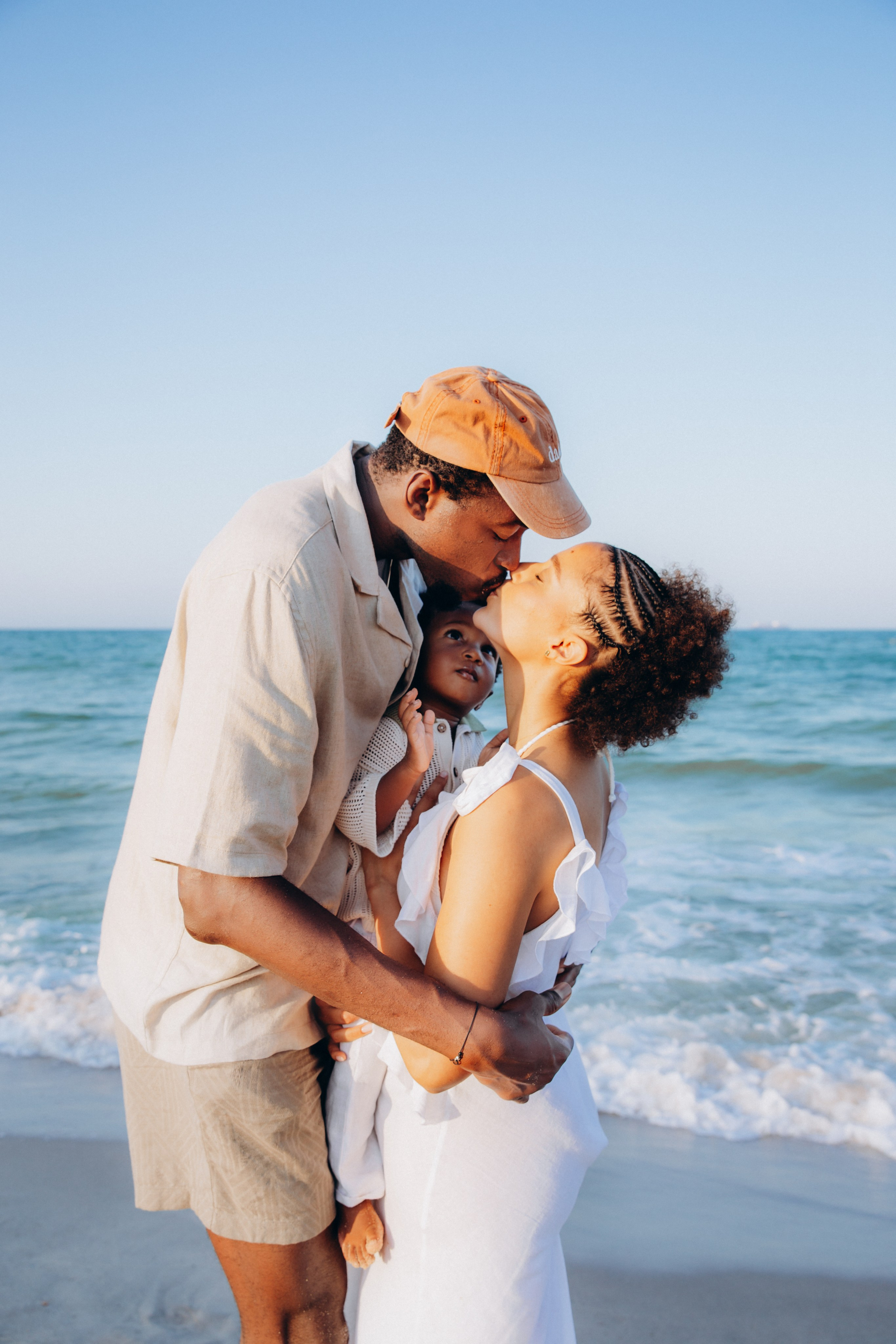 Sesión fotográfica familiar romántica en la playa de Jávea, España — pareja besándose mientras sostienen a su hijo junto al mar, capturando amor, conexión y belleza de la paternidad.