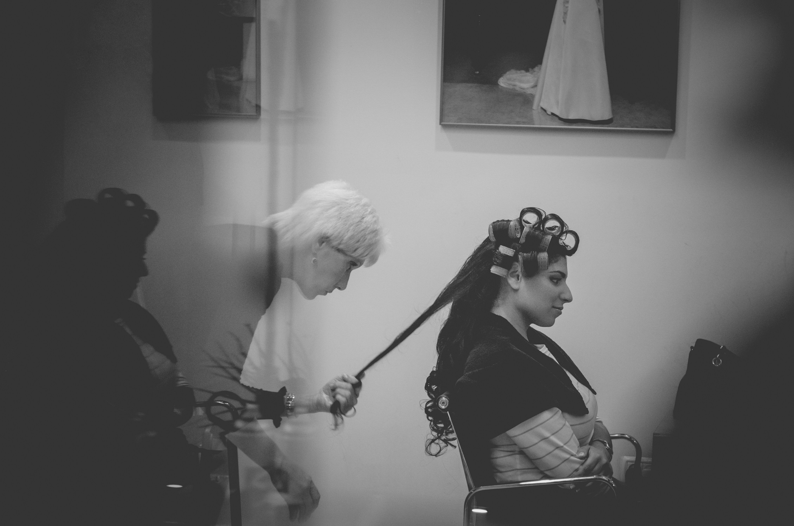 A bride-to-be sits in a salon chair with curlers in her hair while a hairstylist carefully works on her long locks. The black-and-white photograph captures a quiet and reflective moment of wedding preparation, with a soft reflection in the foreground adding depth. A framed picture of a wedding gown hangs on the wall, subtly reinforcing the significance of the day.