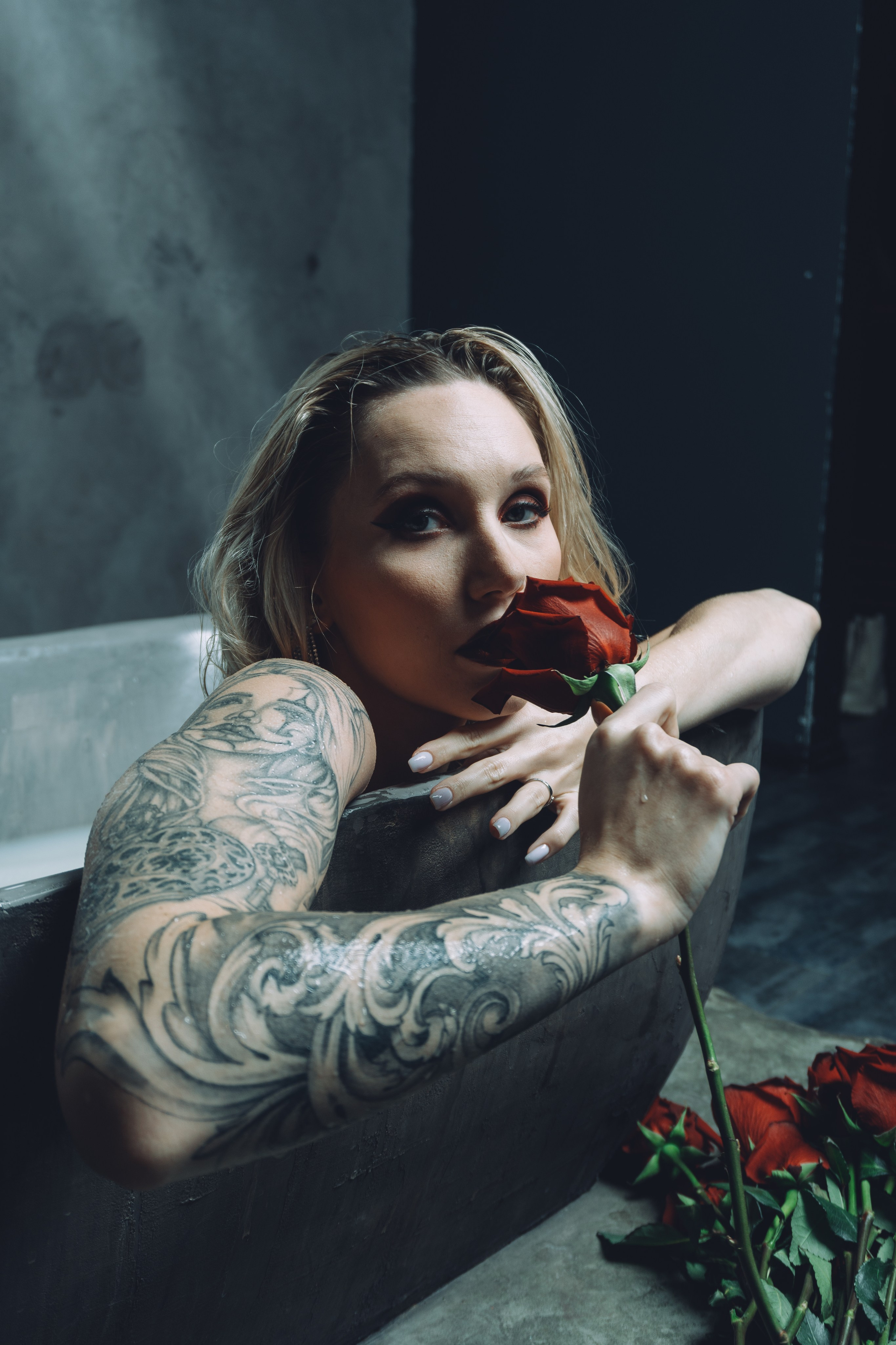 Blonde woman with tattoos smelling a rose in a bath with white water.