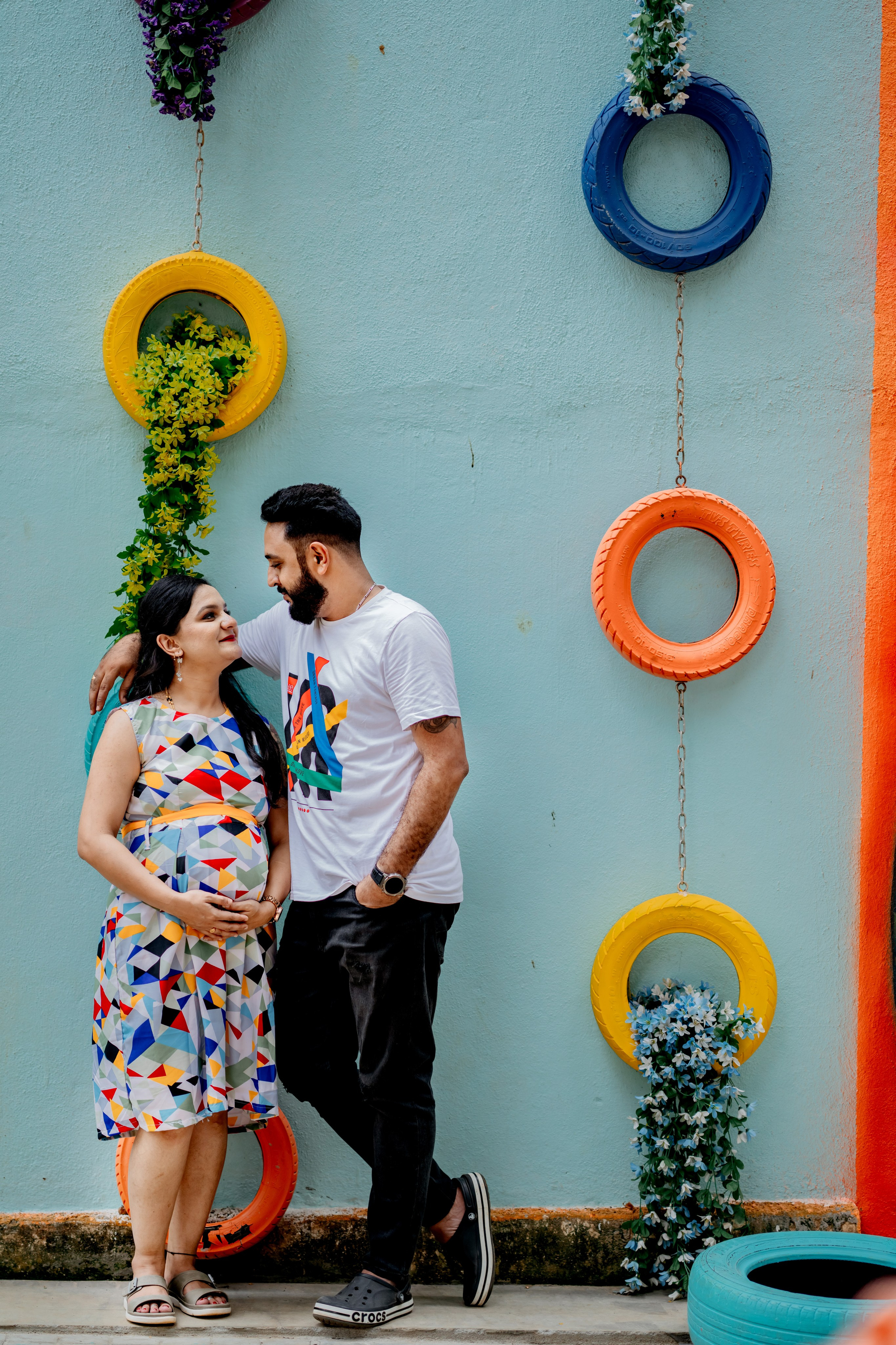 Outdoor maternity photoshoot in Bengaluru featuring a man in a white graphic t-shirt and a woman in a geometric print dress posing against a blue wall with colorful painted tires.