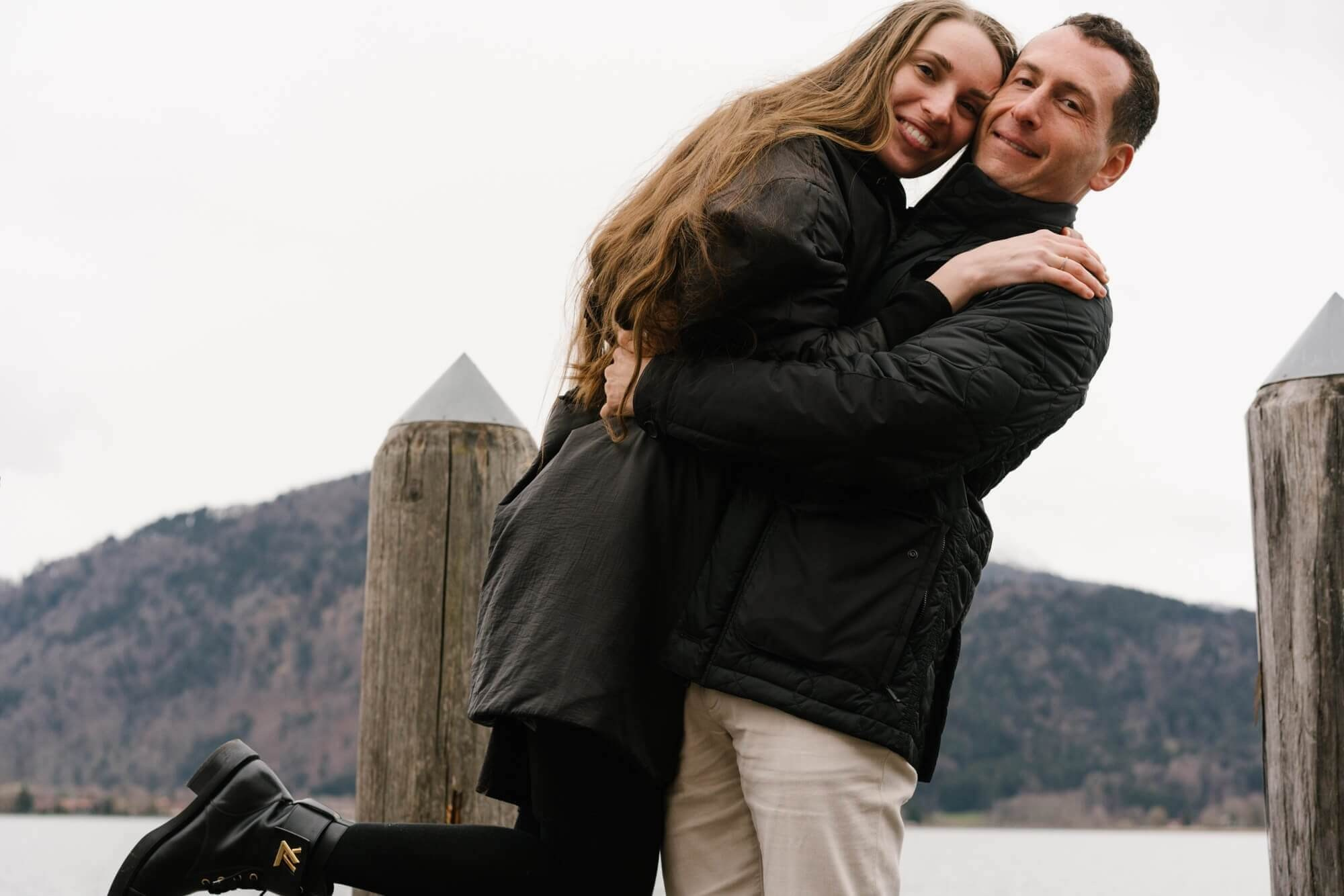 Man carrying woman in his arms on a wooden dock at Tegernsee lake with mountains during couple photo session Bavaria