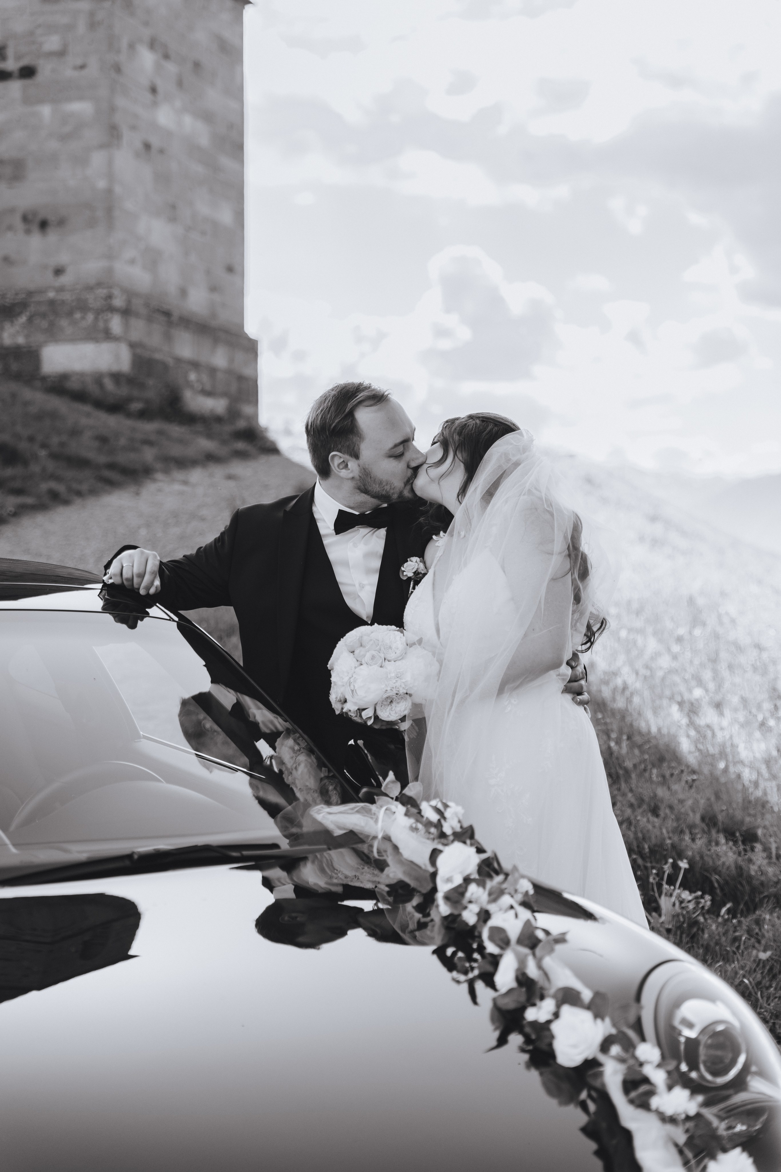 Braut und Bräutigam stehen verliebt am Auto und küssen sich – romantischer Hochzeitsmoment mit stilvollem Flair.
