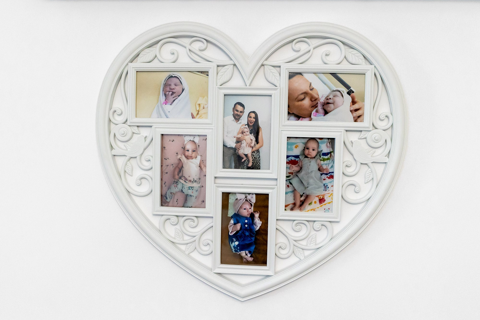 O ramă foto decorativă albă în formă de inimă agățată pe perete, conținând fotografii dragi de familie și imagini cu copii.