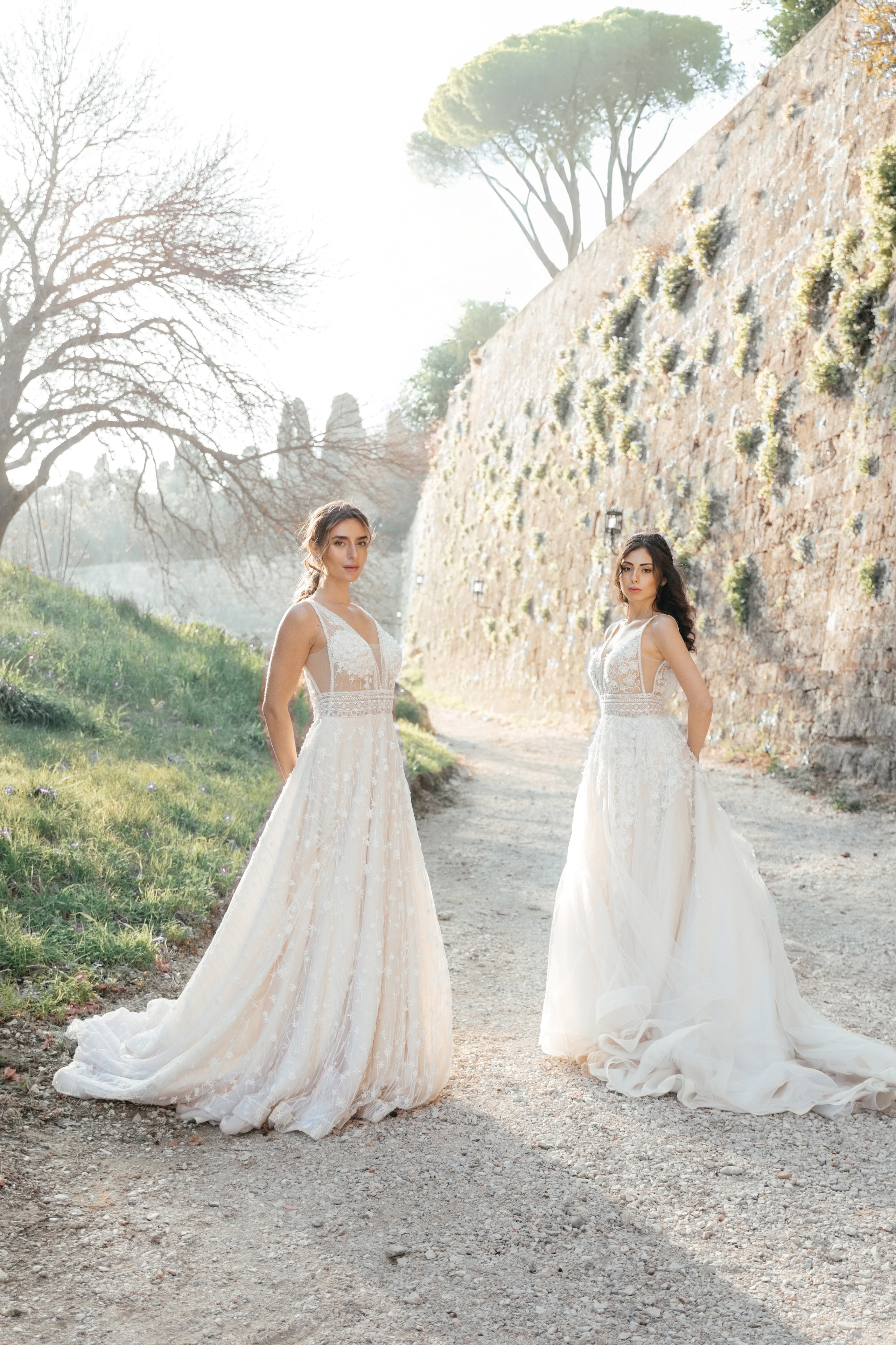 Two brides in elegant wedding dresses pose in the narrow, cobblestone streets of Rhodes' Old Town, surrounded by historic stone buildings and vibrant bougainvillea. The editorial-style photo captures their intimate connection and the timeless charm of the medieval setting, with soft natural light enhancing the romantic atmosphere.