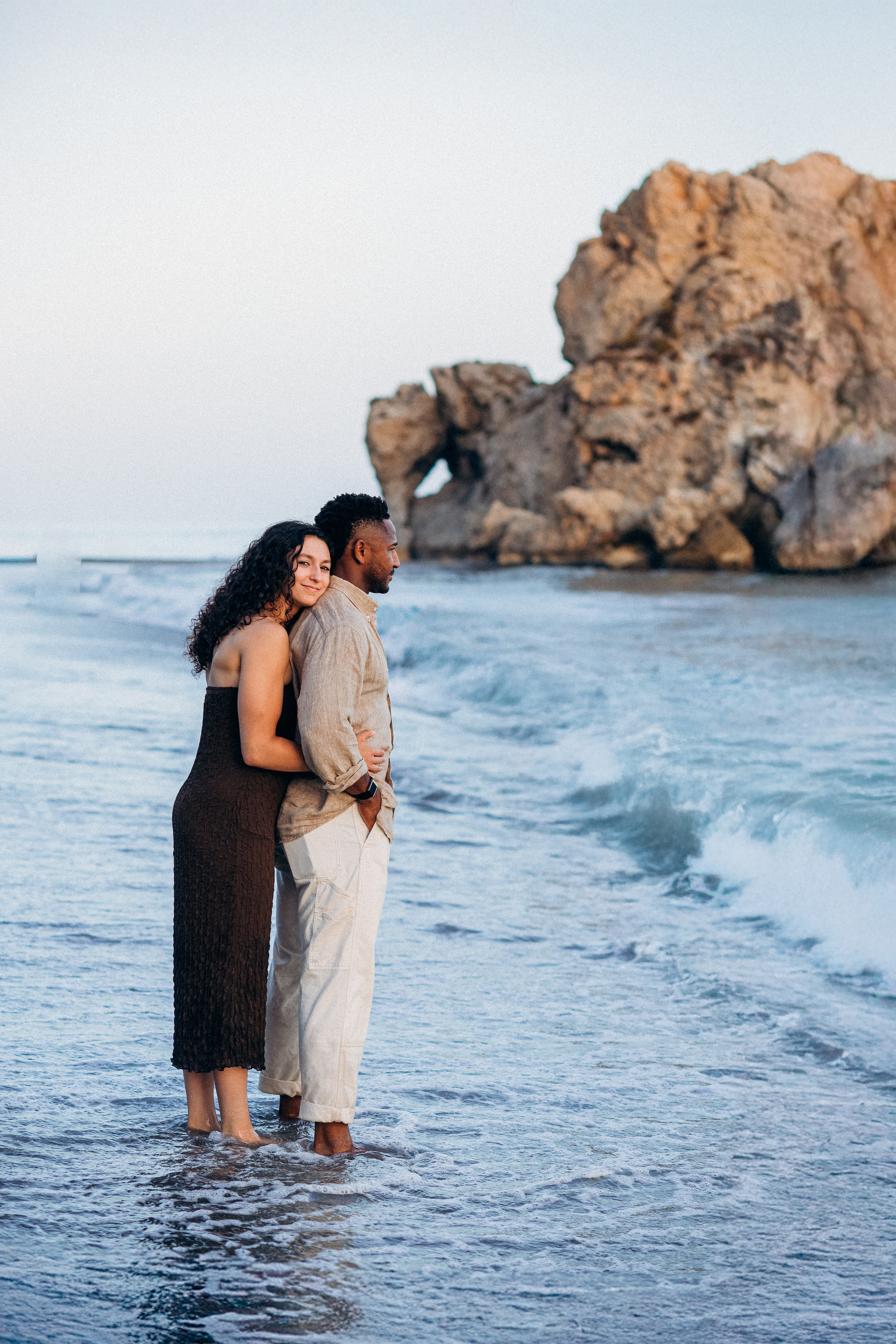 Destination proposal session in Málaga with the newly engaged couple standing barefoot in the sea along Costa del Sol cliffs. Natural light engagement photography highlighting Spain’s dramatic coastline and romantic atmosphere.