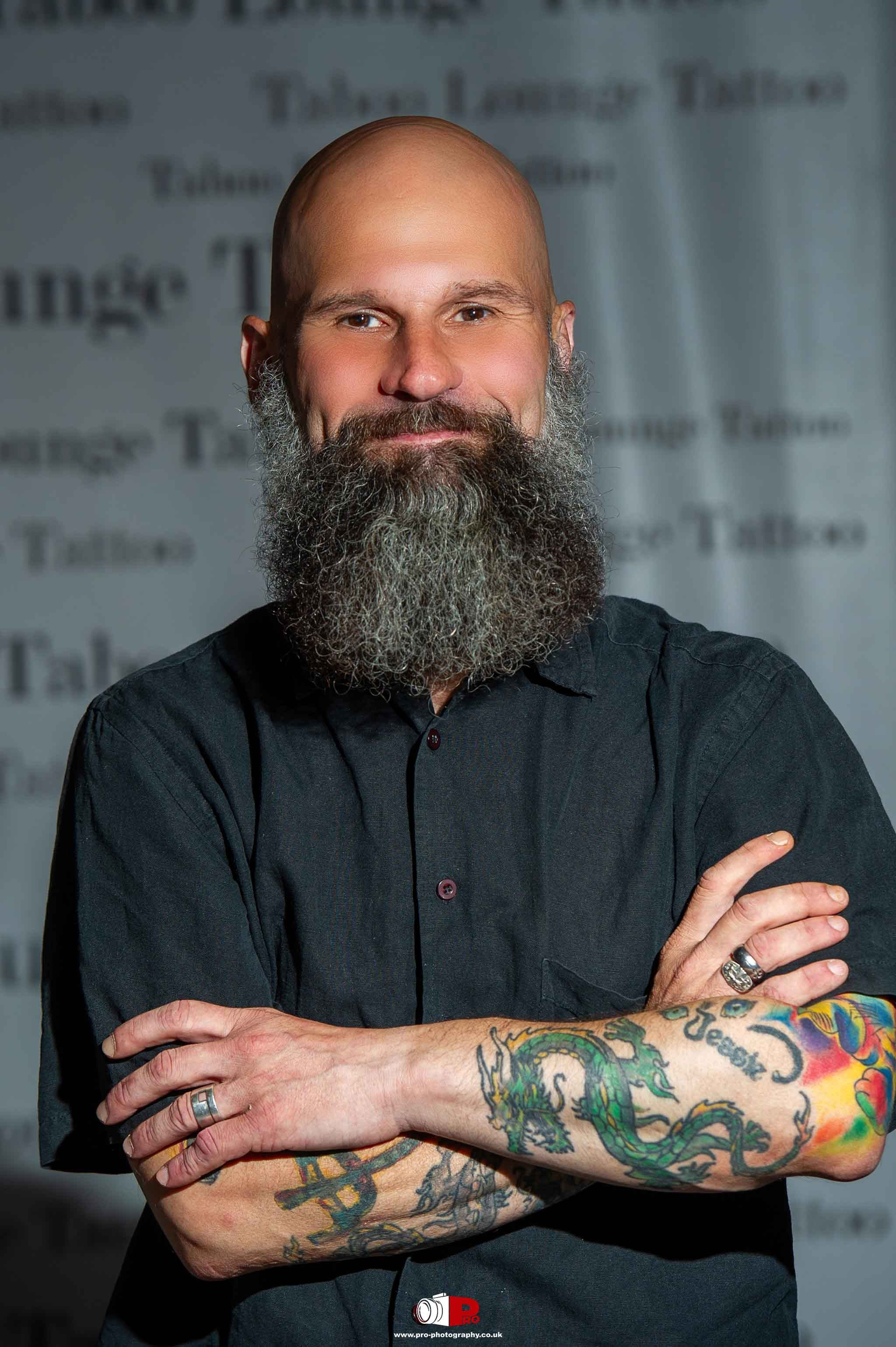 Portrait of a bald, bearded man with colourful tattoos on his arms, wearing a black shirt, smiling confidently in front of a creative background.