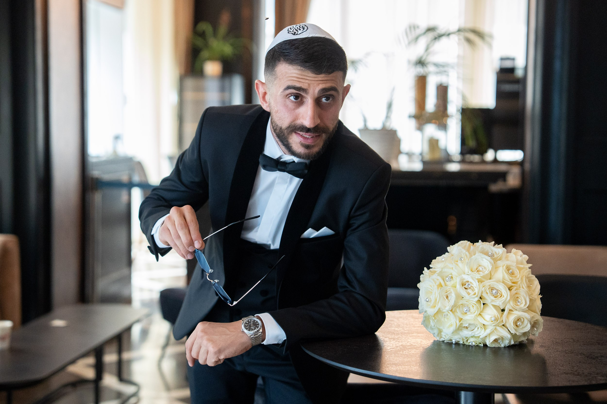 Groom Shimon in a black tuxedo leaning on a table next to a white rose wedding bouquet in Netanya, Israel